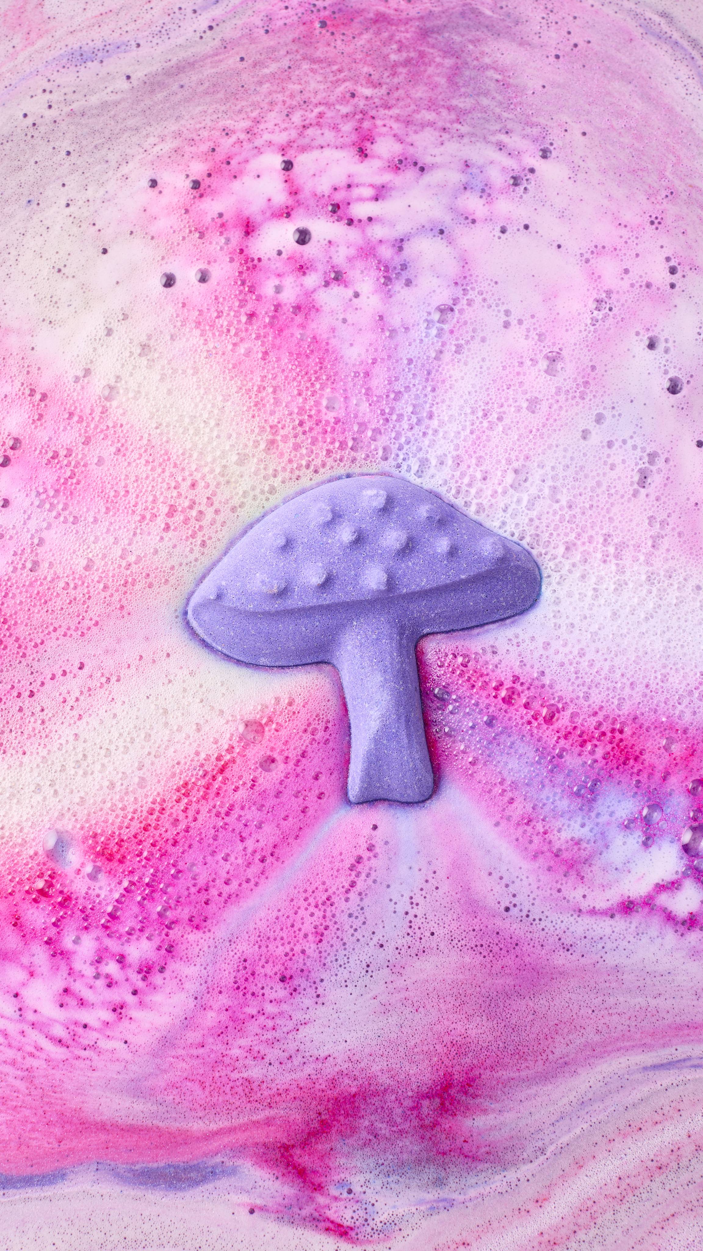 An aerial view shows the "Magik Mushroom bath bomb nearly fully dissolved in water, creating a swirling galaxy of pink, violet, and blue foam. The patterns spiral outwards, leaving a trail of thick, colourful bubbles across the water's surface.