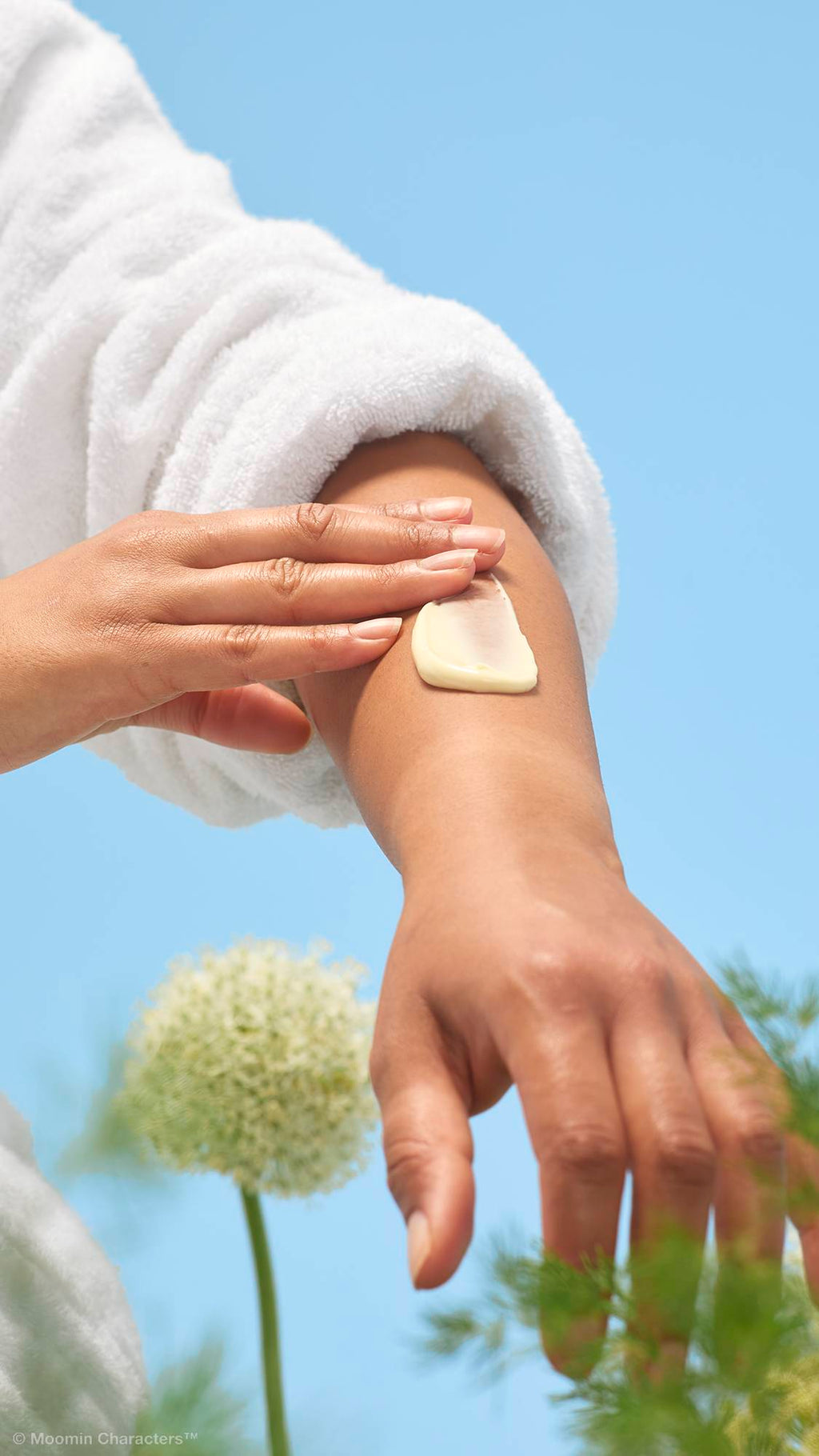 A close-up shot of a person in a white dressing gown smoothing a thick dollop of cream-coloured "Moominmamma" lotion onto their forearm. Soft green foliage and a white flower are visible in the background against a clear blue sky.