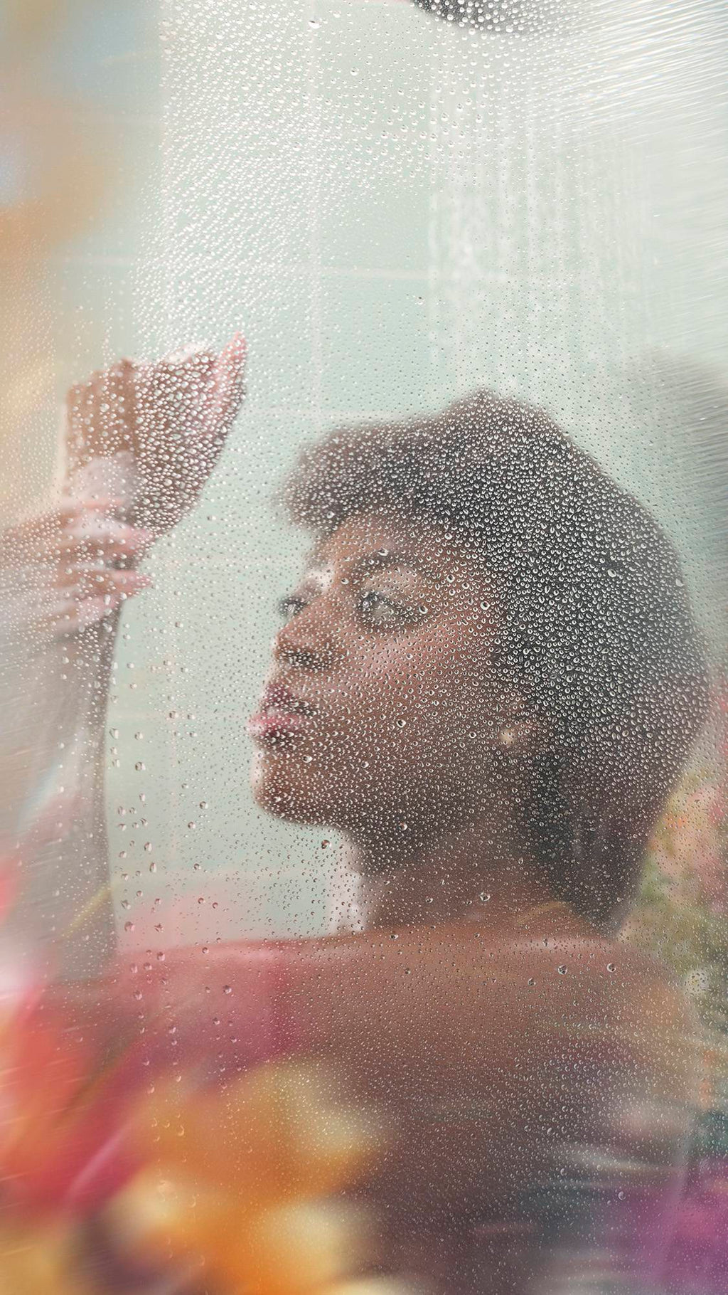 A person with short, dark curly hair stands behind a wet shower screen covered in water droplets. They are looking upwards with one hand raised near their face, set against a backdrop of mint green tiles and blurred colorful flowers.