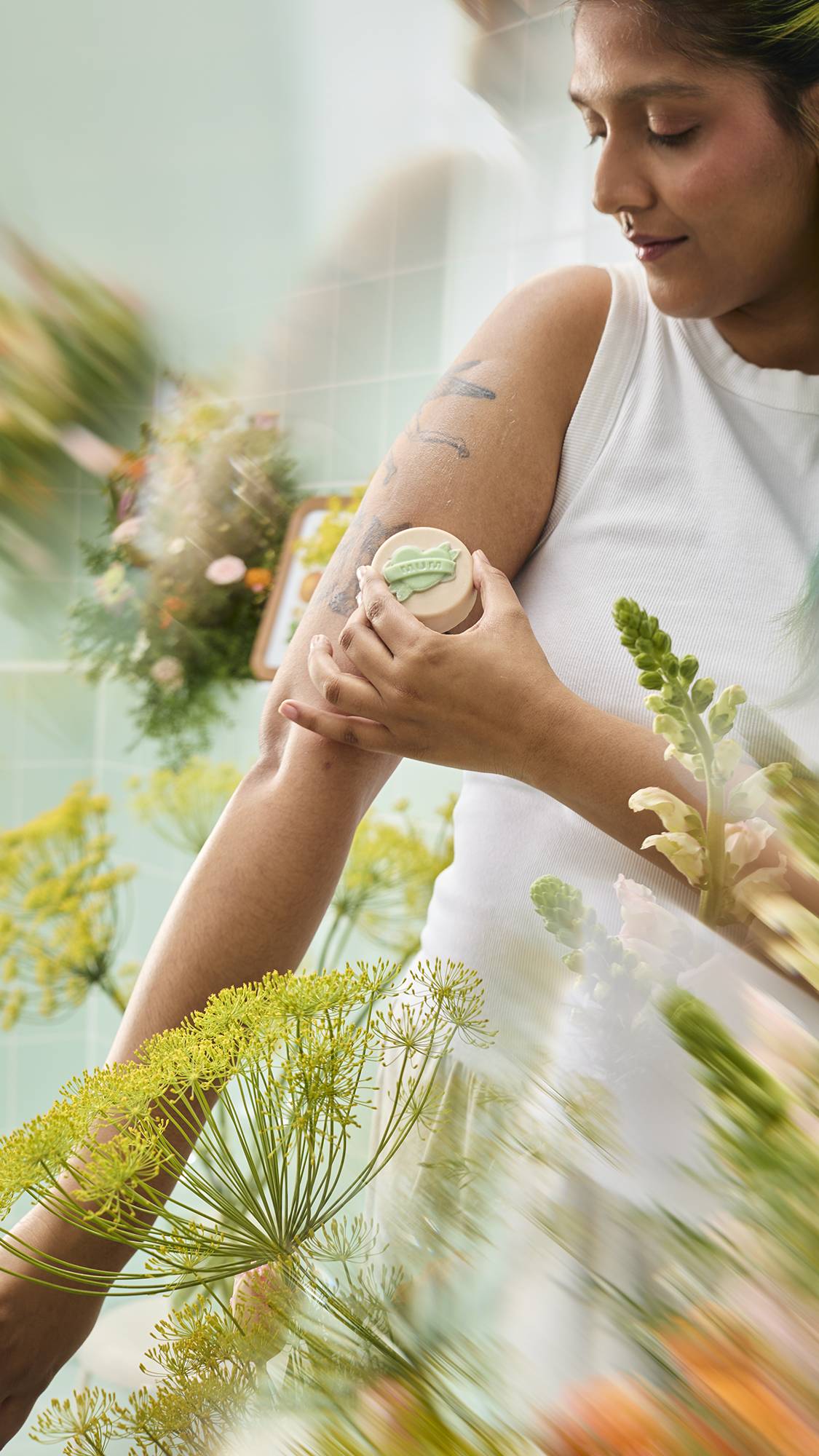 A person wearing a white tank top applies a round, cream-coloured "Symbol of Love" body oil bar to their tattooed upper arm. The scene is framed by blurred yellow flowers against a background of mint green tiles.