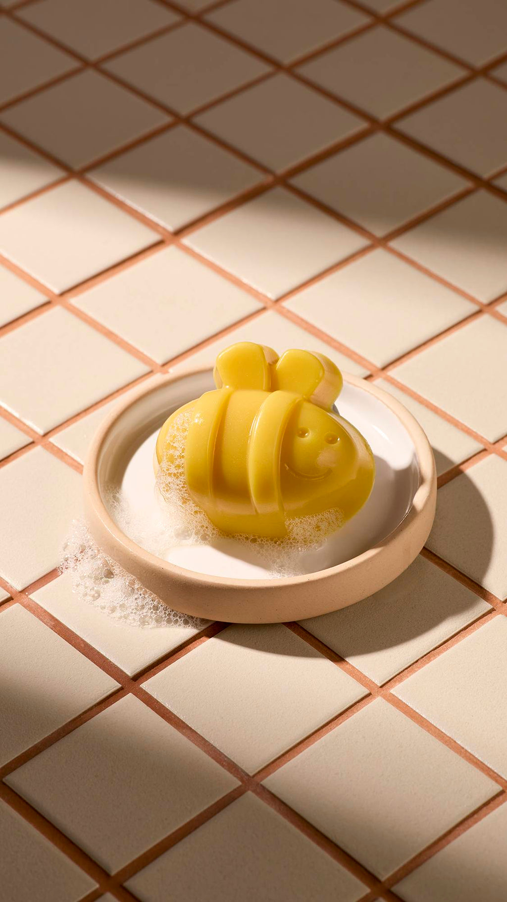 A yellow bee-shaped facial gummy wash with a smiling face rests in a cream-coloured dish. The soap is covered in light white bubbles, and the dish sits on a background of beige square tiles under soft sunlight and shadows.