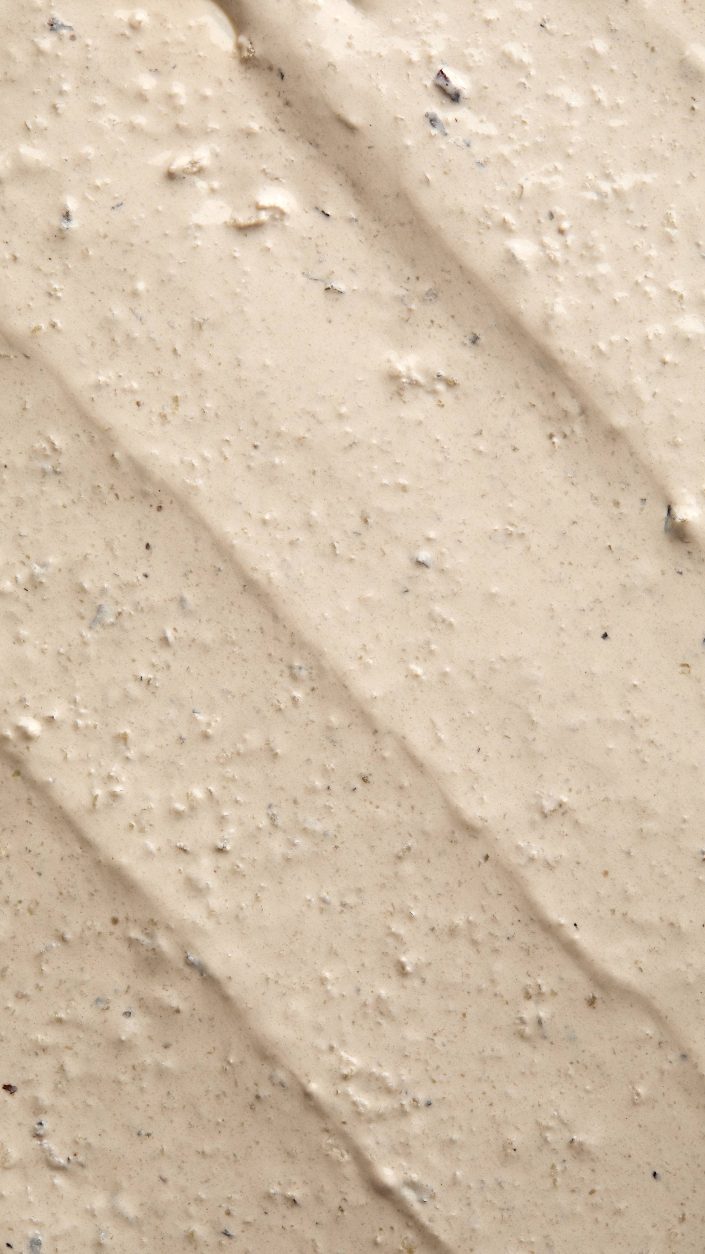 A close-up, textured shot of "Tiny Musicians Of The Forest, a creamy, beige-coloured face mask spread in thick, diagonal ridges. The surface is grainy and lumpy, containing small dark exfoliating particles and flecks.