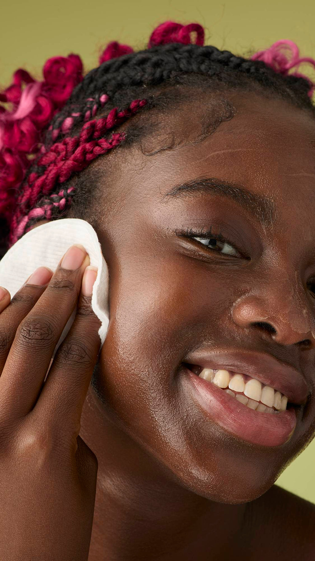 A person with pink braids smiles while gently massaging the melted cleanser onto their jawline and cheeks against an olive green background.
