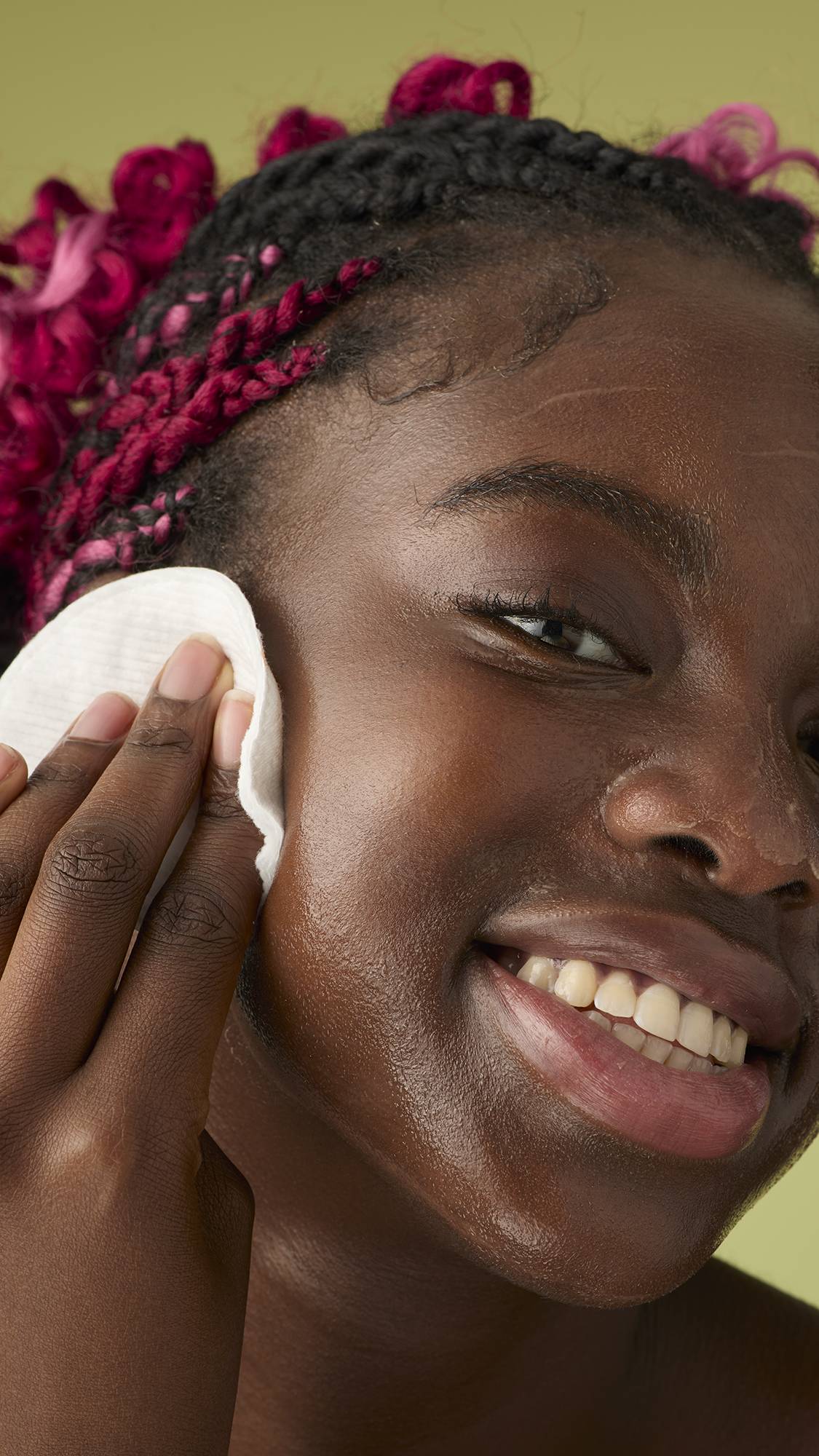 A person with pink braids smiles while gently massaging the melted cleanser onto their jawline and cheeks against an olive green background.