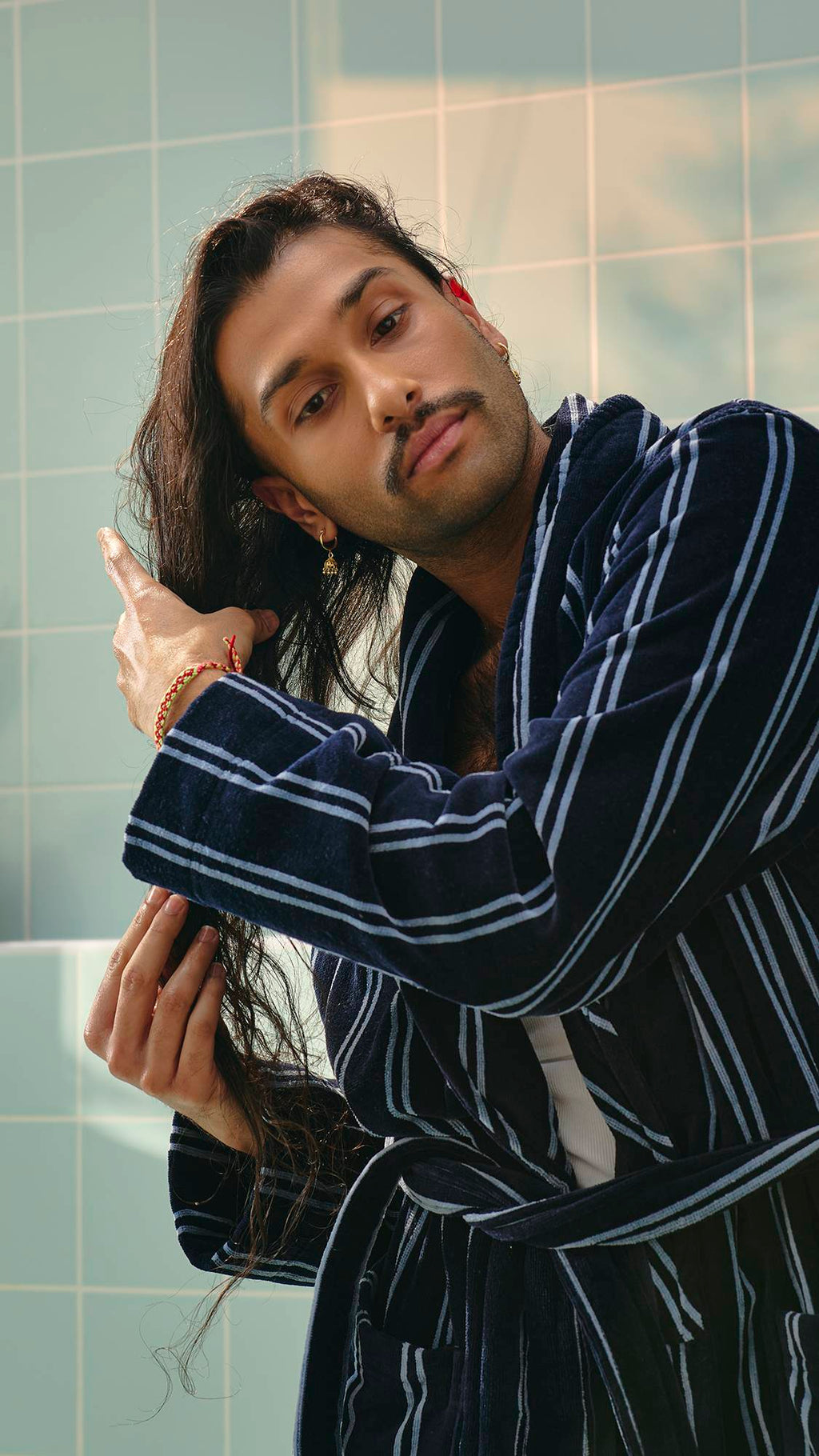 A model with long dark hair and a moustache, wearing a navy and white striped dressing gown, runs their hands through their hair to apply the "Vegan Leather Jacket" hair and beard oil. Light green tiles are visible in the background.