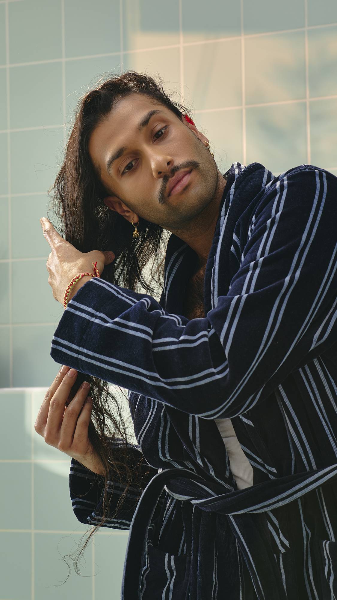 A model with long dark hair and a moustache, wearing a navy and white striped dressing gown, runs their hands through their hair to apply the "Vegan Leather Jacket" hair and beard oil. Light green tiles are visible in the background.