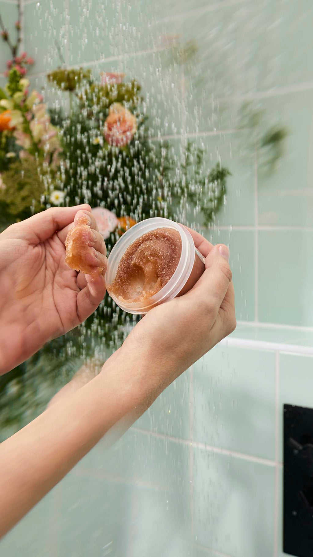 Two wet hands hold an open pot of "Crème Brûlée" Body Scrub under a shower stream against a background of green tiles and flowers. One hand displays a scooped chunk of the thick, golden-brown granular scrub to show its texture.