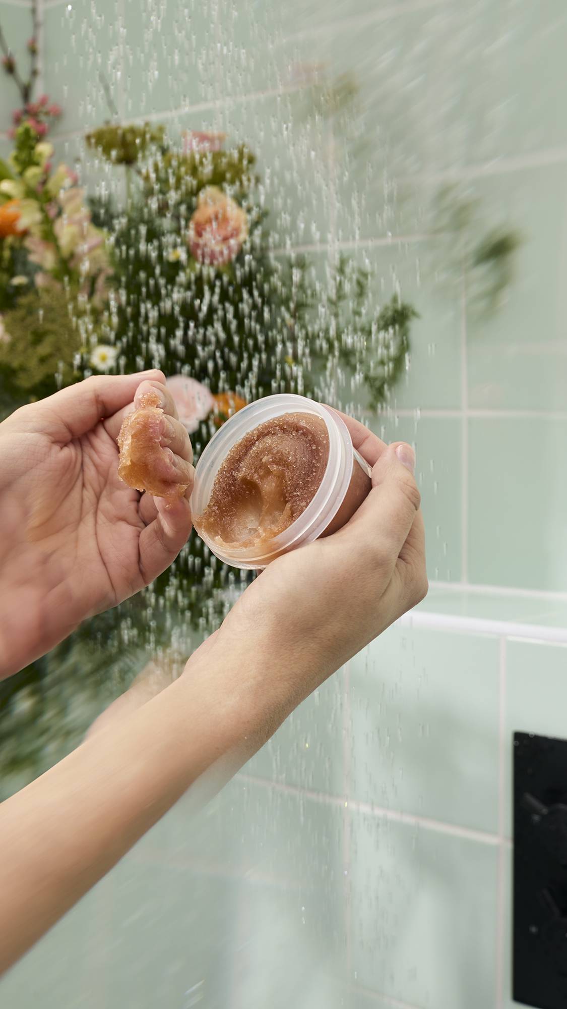 Two wet hands hold an open pot of "Crème Brûlée" Body Scrub under a shower stream against a background of green tiles and flowers. One hand displays a scooped chunk of the thick, golden-brown granular scrub to show its texture.