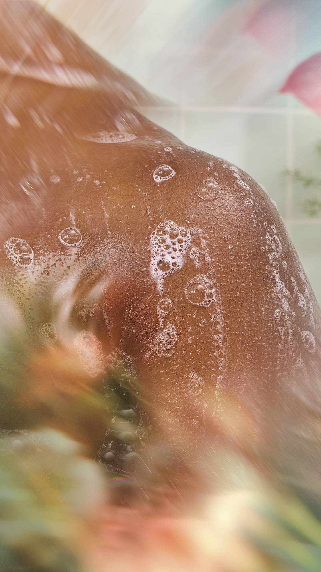 A close-up shows water streaming down a wet shoulder, with small soapy bubbles clinging to the skin. The scene is framed by blurred pink and green floral elements against a mint green tiled wall.