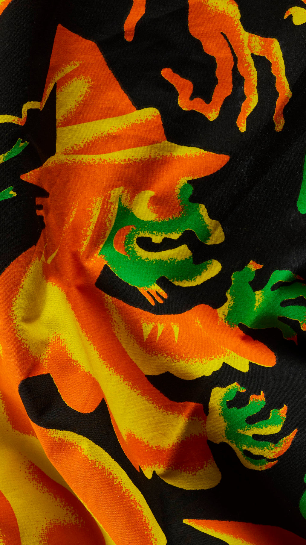 A close-up of a rumpled black fabric knot wrap with a vibrant Halloween illustration. It depicts a cackling witch with a bright orange pointy hat and cloak, a green face, and outspread green hands outlined in yellow and orange.