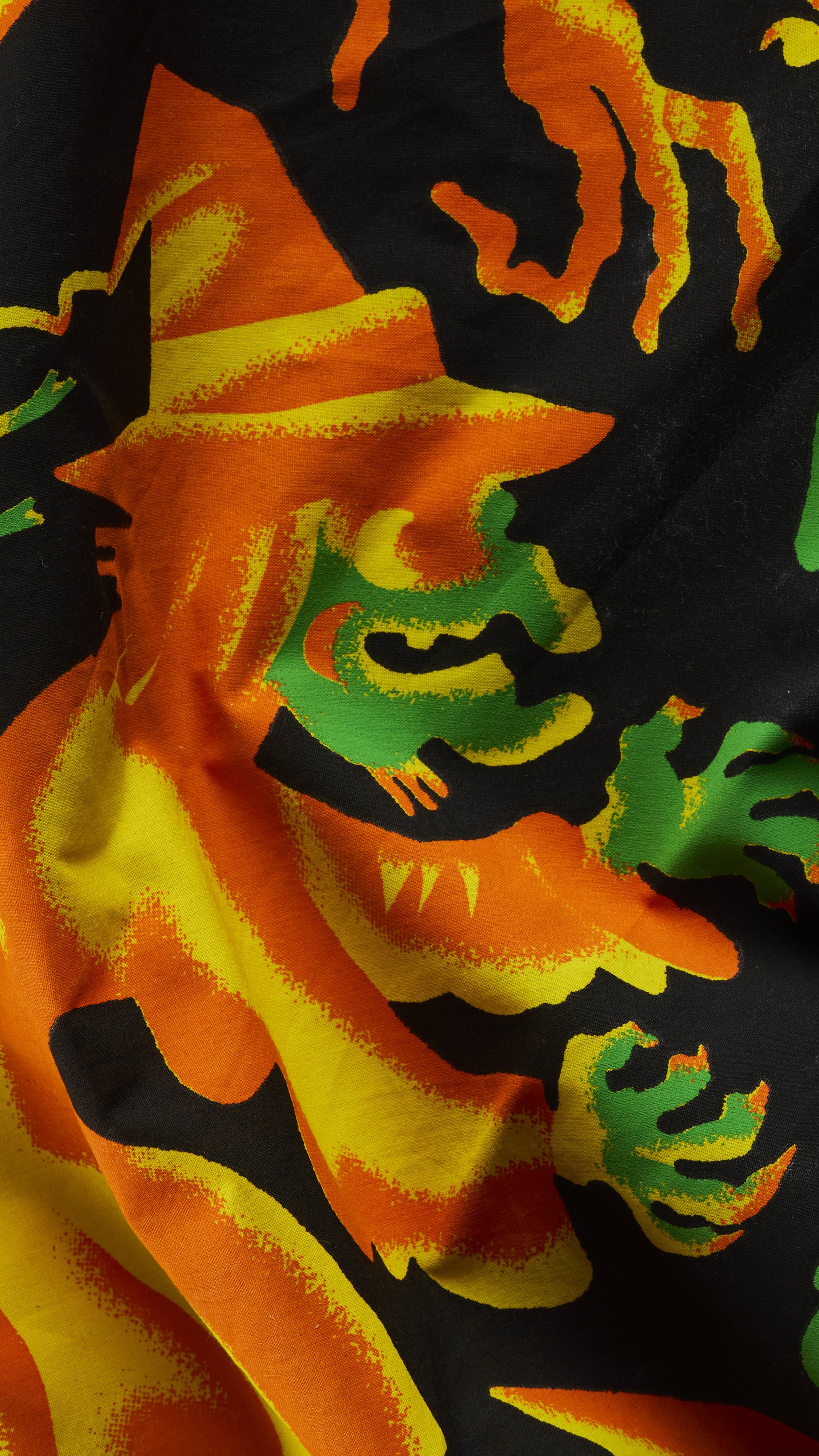 A close-up of a rumpled black fabric knot wrap with a vibrant Halloween illustration. It depicts a cackling witch with a bright orange pointy hat and cloak, a green face, and outspread green hands outlined in yellow and orange.