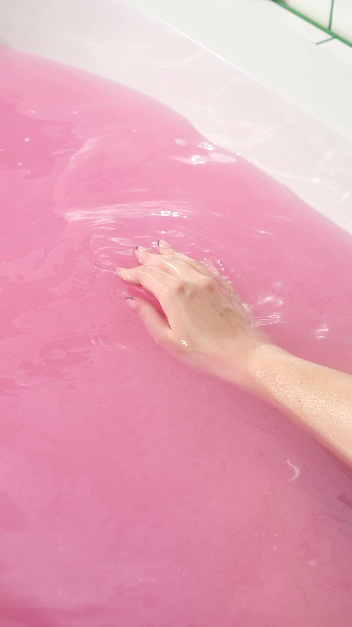 The Sex Bomb Bath Bomb has completely dissolved, leaving the bathtub filled with a vibrant, opaque shade of deep pink. A hand is shown gently submerged in the colorful water, illustrating the relaxing and aesthetically pleasing result of the bath.