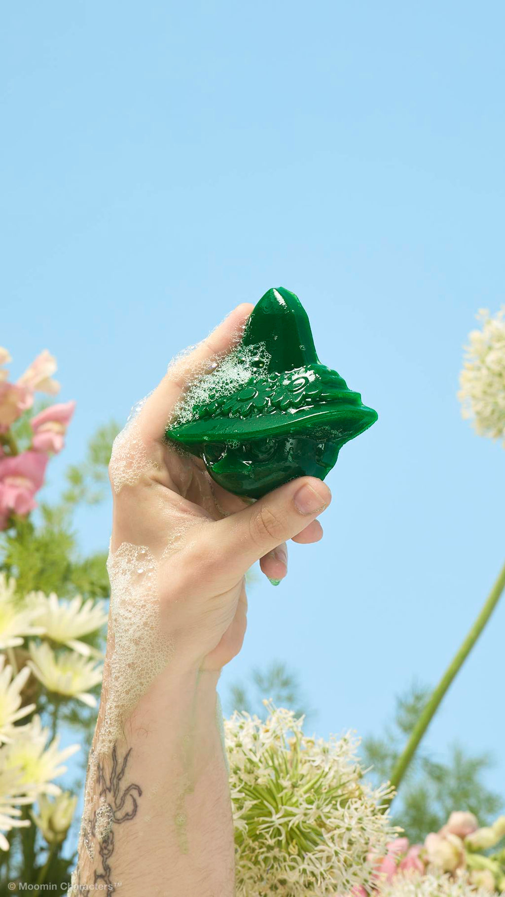 A close-up shot of a tattooed arm holding up a green, soapy Snufkin-shaped shower gummy covered in frothy bubbles. Yellow, white, and pink wildflowers are visible in the background against a clear blue sky.