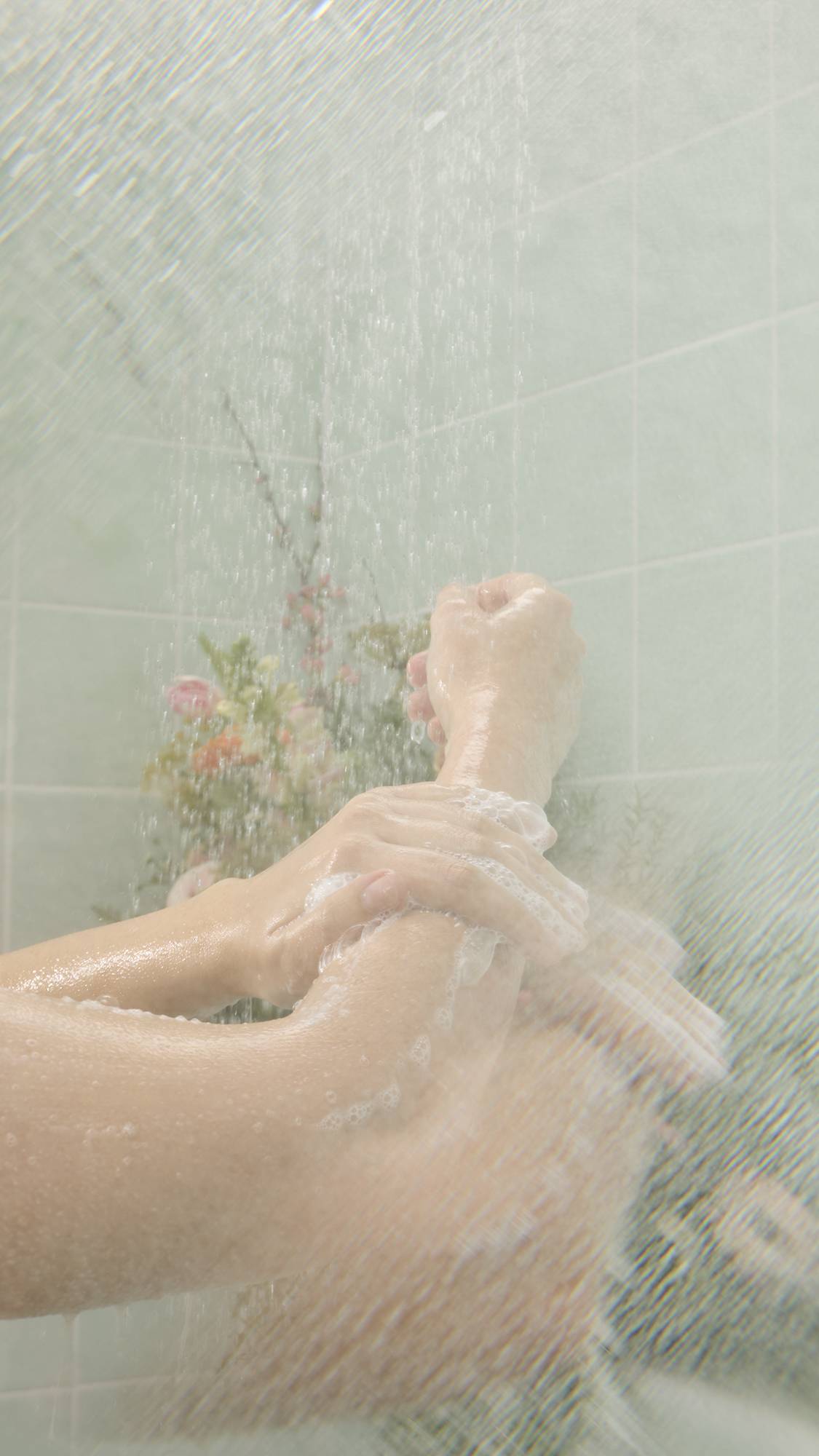 Wet, soapy hands massage "Crème Brûlée" Body Scrub onto an arm under a shower stream. The lathering action takes place against a background of mint green tiles and soft-focus flowers.