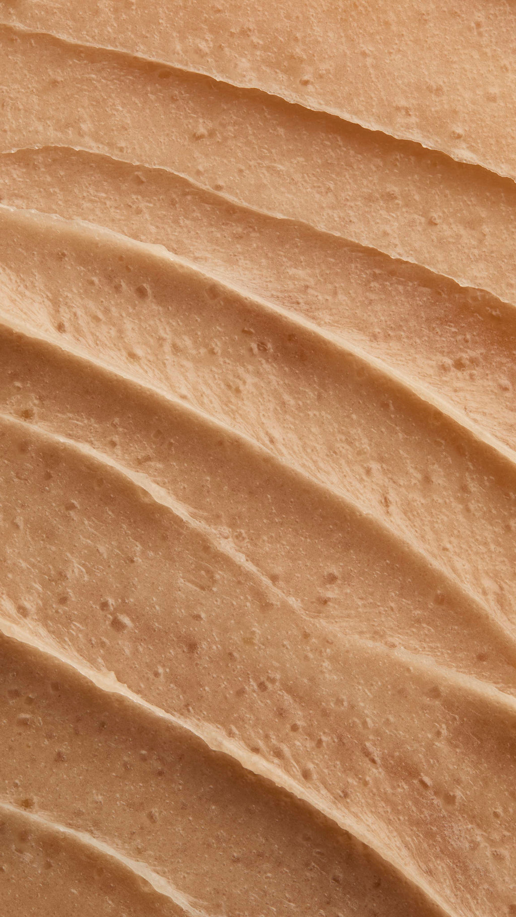 A close-up, full-frame shot displays the texture of the "Crème Brûlée" Body Scrub. The creamy, light brown mixture is spread into thick, wavy ridges, with small exfoliating granules visible throughout the paste.