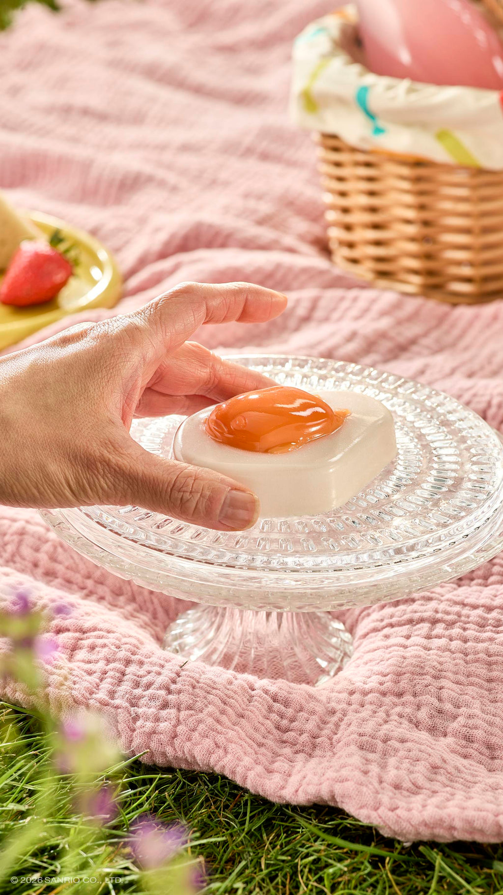 A hand reaches for a glossy fried-egg-shaped shower jelly sitting on a glass cake stand. The sunny picnic scene features a pink blanket on the grass, a wicker basket, and a strawberry.