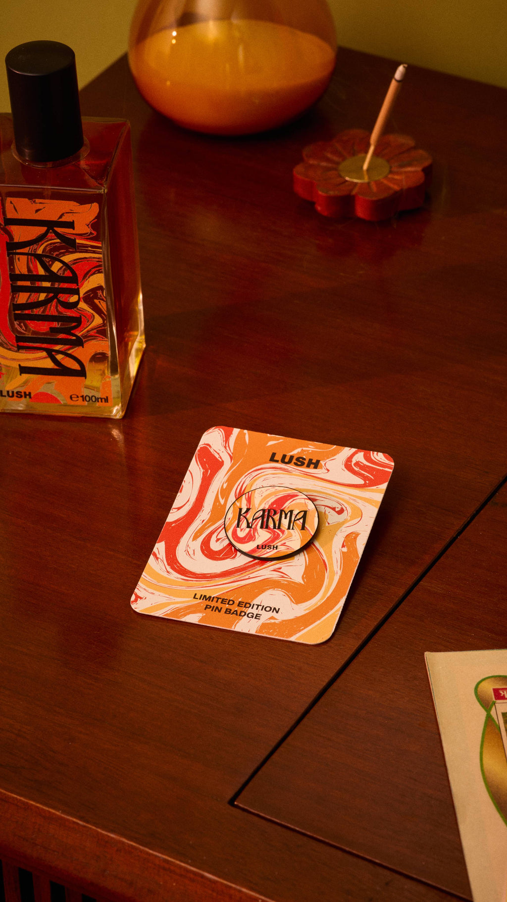 The image shows a Karma pin badge on its square backing card, featuring a red, orange, and white marbled design, resting on a wooden surface. A Karma perfume bottle and other items sit blurred in the background.
