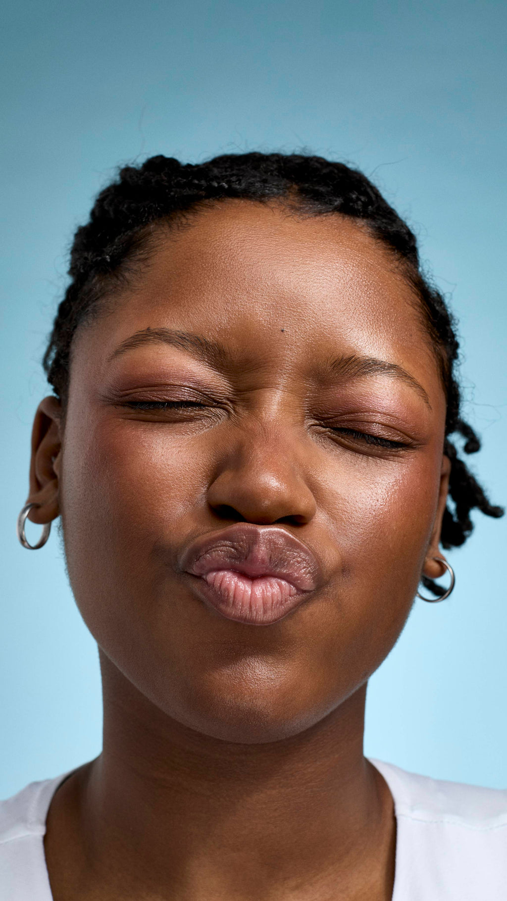 A close-up of a model with braided hair and glowing skin, making a playful kissing face with their eyes closed. They are wearing a white top and small silver hoop earrings against a soft, light-blue background.
