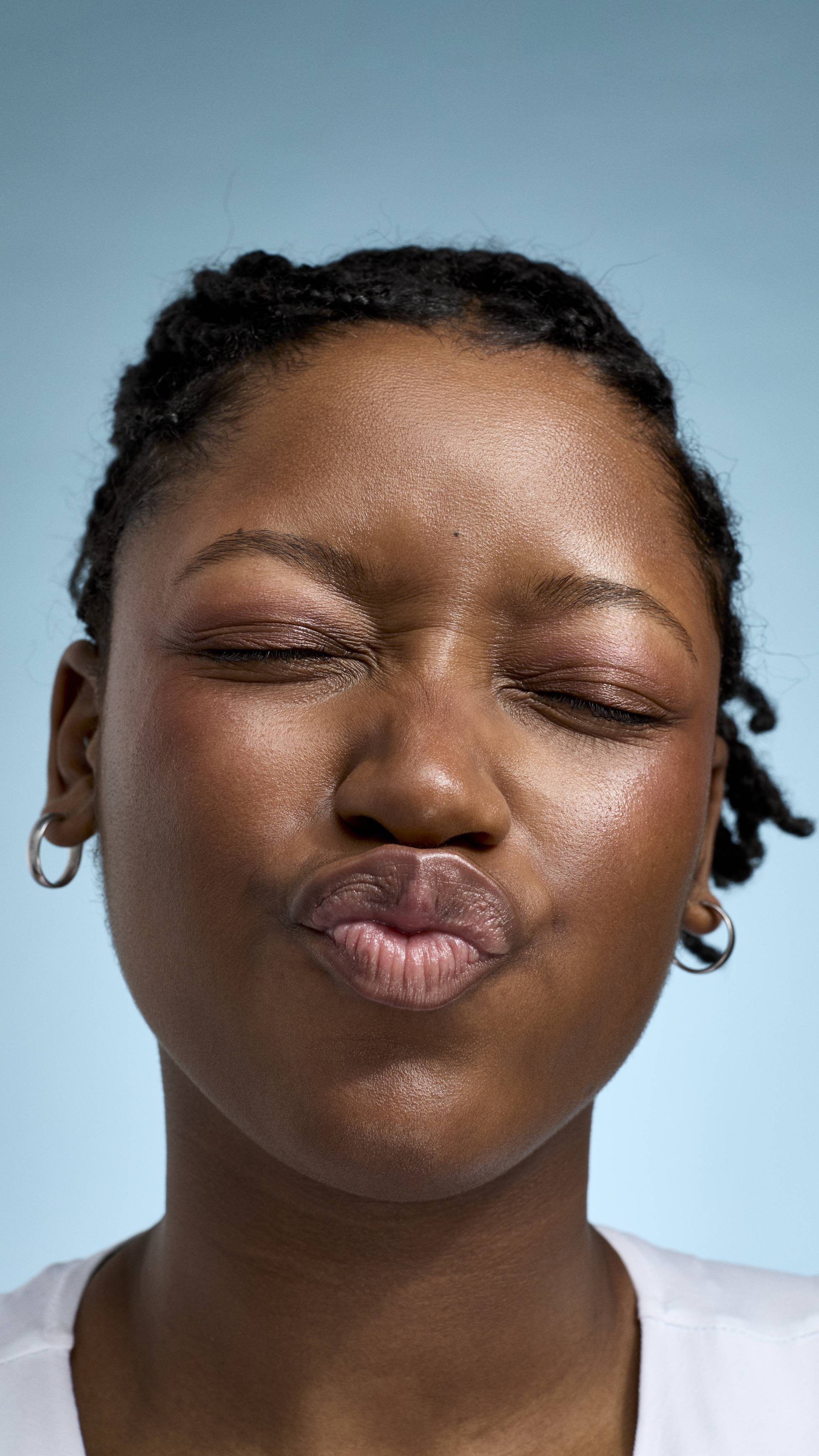 A close-up of a model with braided hair and glowing skin, making a playful kissing face with their eyes closed. They are wearing a white top and small silver hoop earrings against a soft, light-blue background.