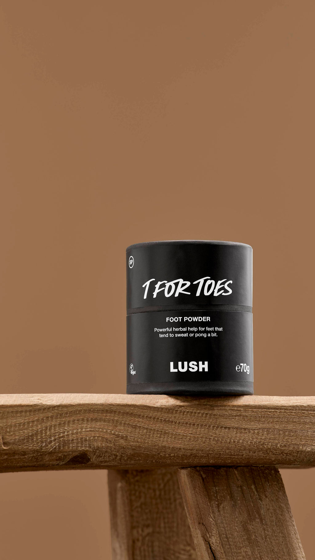 The image shows a black, cylindrical, cardboard tub of 'T For Toes' foot powder with a black lid. The tub has white text on its label, and it stands upright on a rustic wooden surface against a brown background.