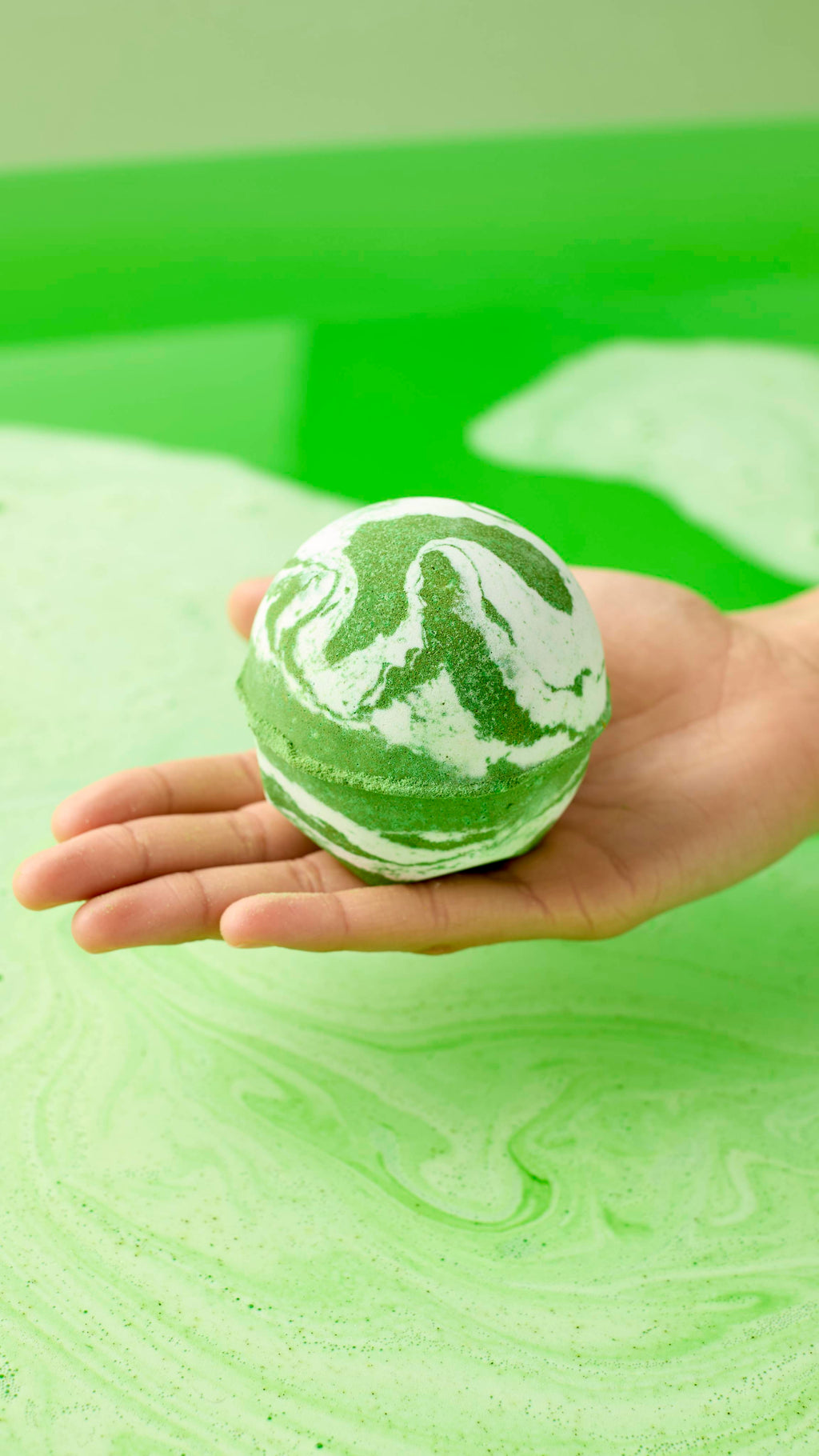 A hand holds the green and white marbled Cold Bath Bomb over a bathtub filled with vibrant green water. The bathwater swirls with light green foam, perfectly matching the colors of the bath bomb.