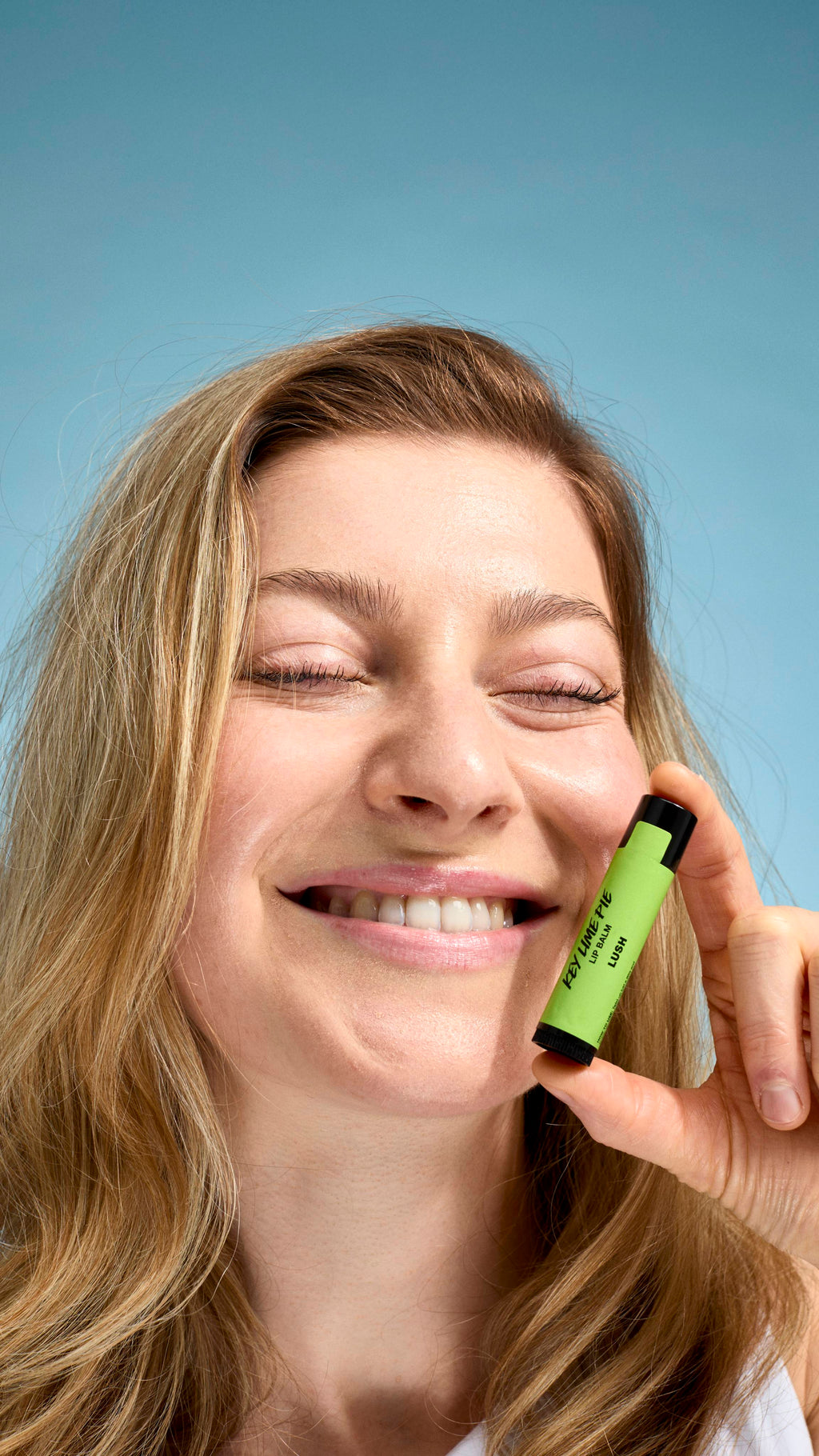 A close-up of a model with fair skin and dark blonde hair, smiling with their eyes closed. They are holding a bright green-labelled tube of Lush 'Key Lime Pie' lip balm against their cheek. The background is a soft, light blue.