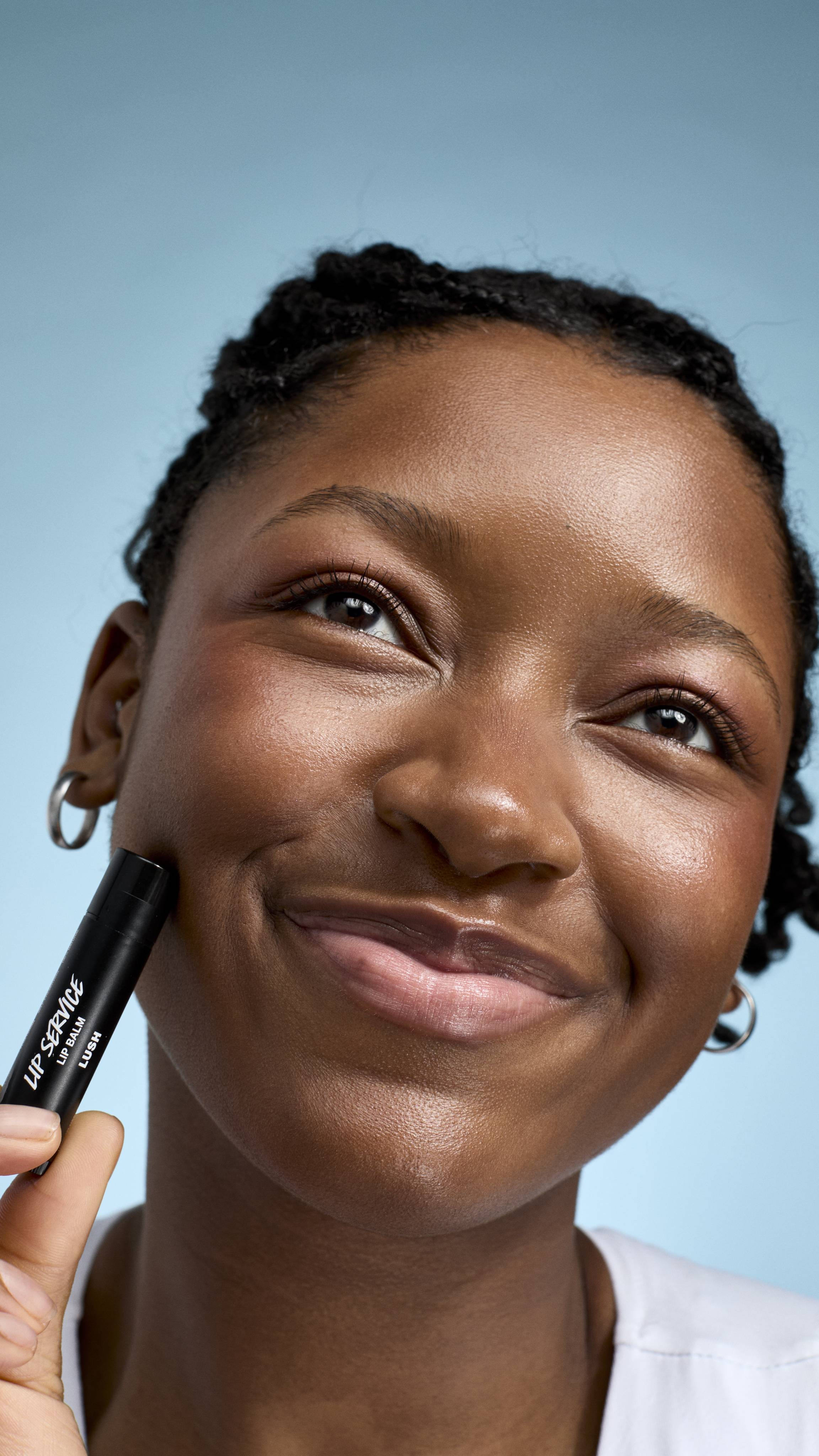 A close-up of a model with braided hair and glowing skin smiling warmly. They hold a black tube of Lush 'Lip Service' lip balm against their cheek while looking slightly off-camera against a soft blue background.