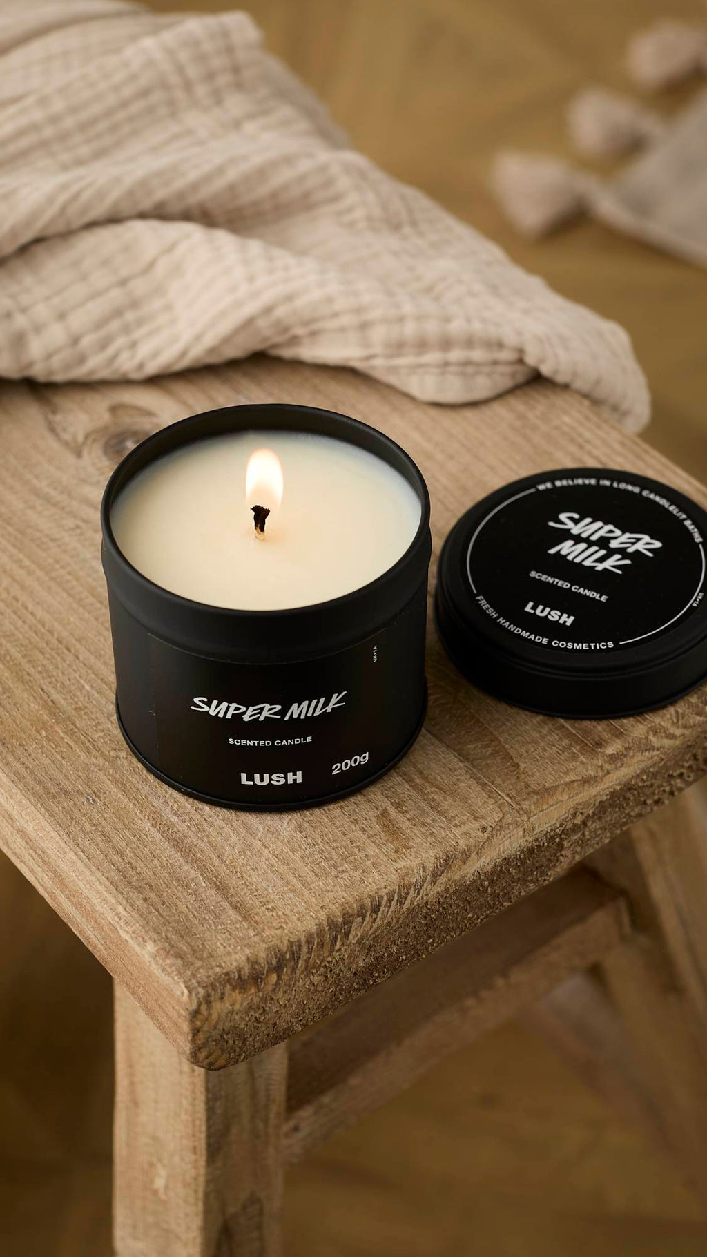 A close-up, slightly aerial view of the black Super Milk Scented Candle lit on a weathered wooden stool. The creamy white wax is melting around the bright wick, with the lid resting beside it and a beige cloth draped in the background.