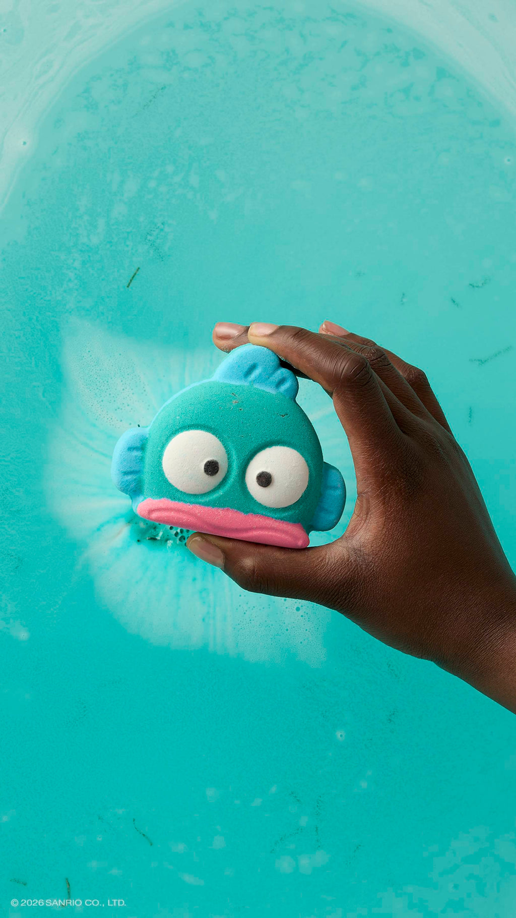 A hand holds a teal, fish-shaped bath bomb over a bath of bright turquoise water. The bath bomb features a pink mouth and large, startled white eyes. A soft white fizz is visible in the water directly behind the product.