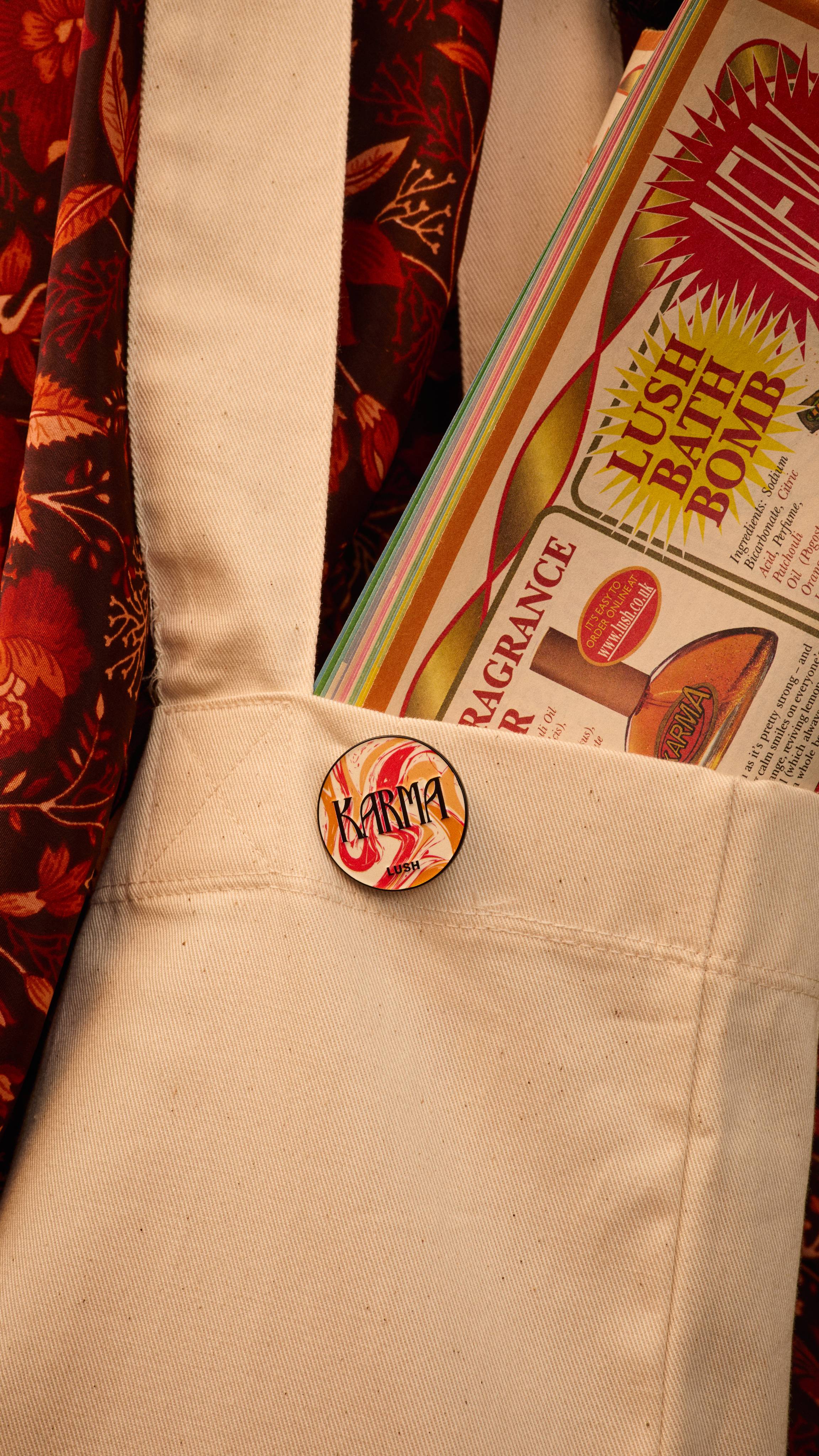 The image shows the red, orange and white marbled design Karma pin badge attached to a light canvas tote bag pocket. A colourful Lush magazine is visible inside, with a patterned fabric backdrop.