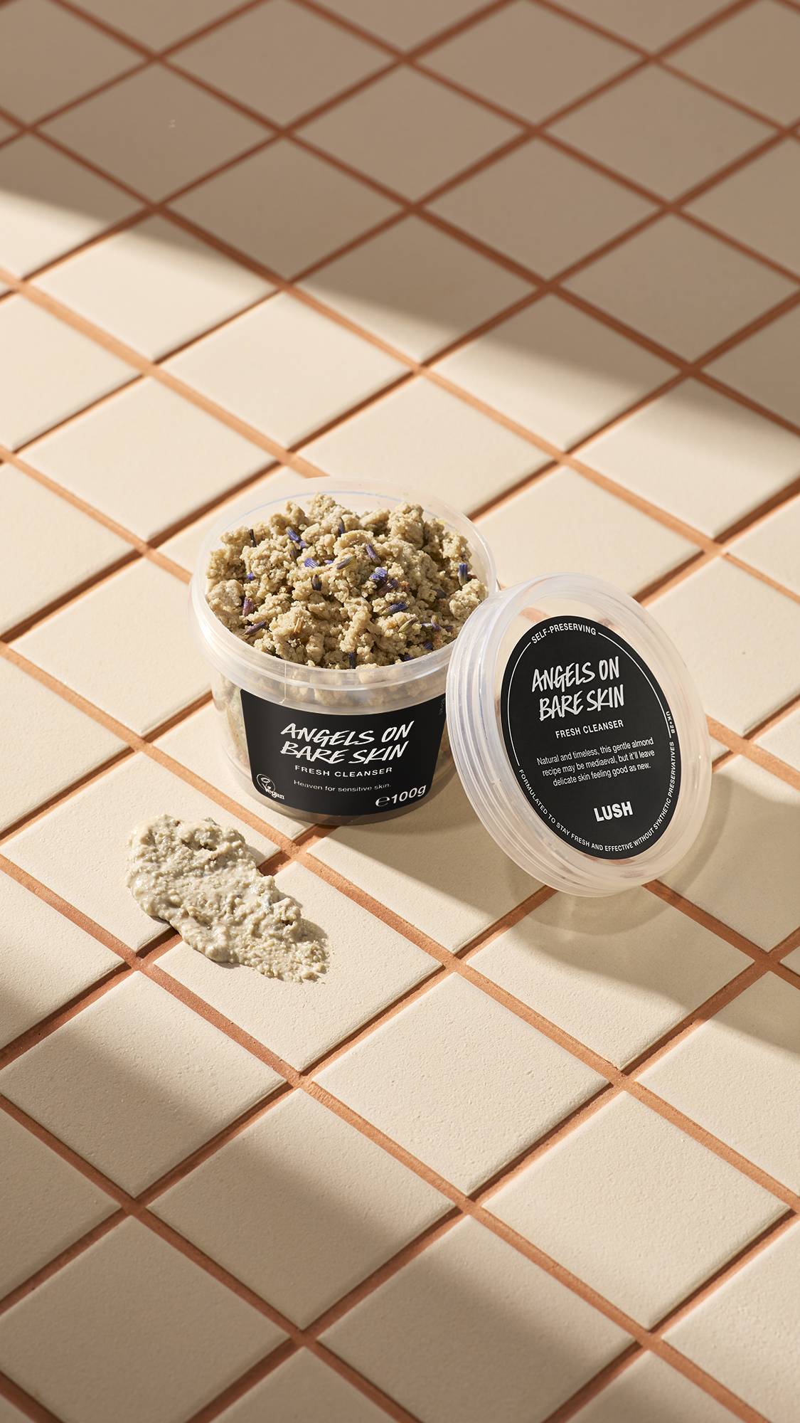 An open clear pot of Lush Angels on Bare Skin Fresh Cleanser, showing a crumbly lavender-flecked paste, sits on beige tiles. Its lid rests alongside it next to a smear of the creamy mixture, illuminated by soft diagonal sunlight.