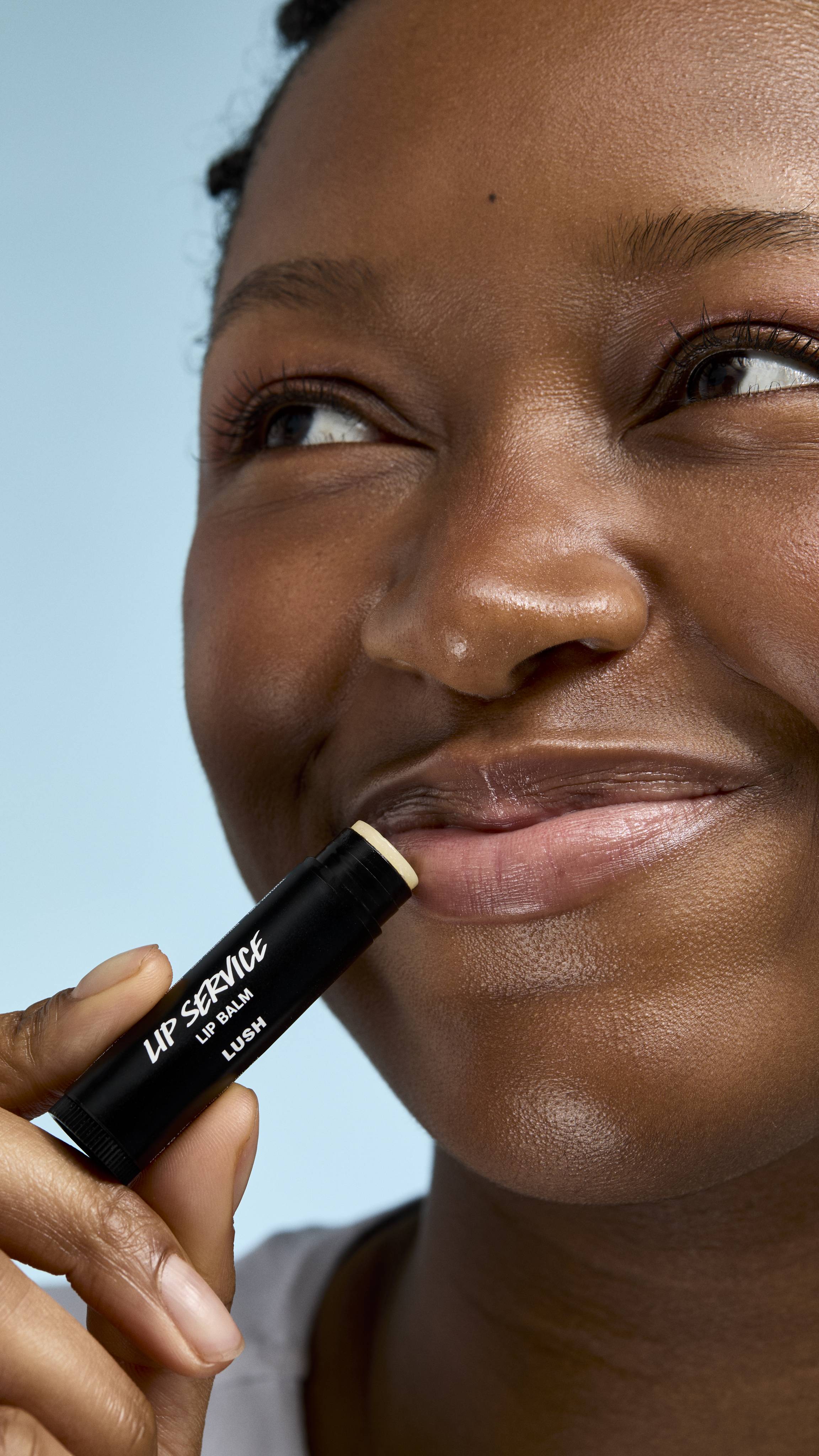 A close-up of a model applying 'Lip Service' lip balm to their lips. They have a soft smile and are looking slightly off-camera, holding the black Lush tube against their mouth against a clear, light-blue background.