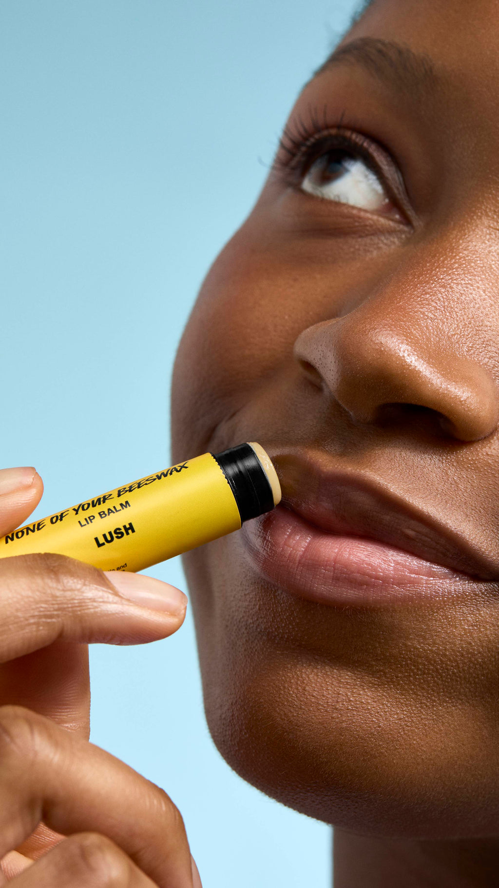 A close-up of a model applying 'None of Your Beeswax' lip balm to their lips. They are looking upwards with a soft smile, holding the bright yellow Lush tube against their mouth. The background is a clear, light blue.