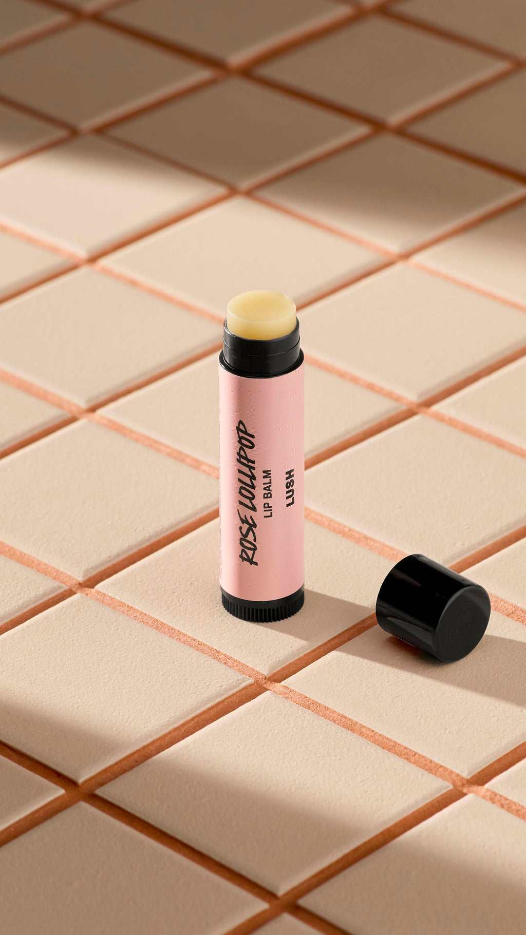 A light pink-labelled tube of Lush 'Rose Lollipop' lip balm stands upright on light-coloured square tiles. The black cap is off, lying next to the base, revealing the pale, creamy balm inside. Soft shadows fall across the tiled surface.