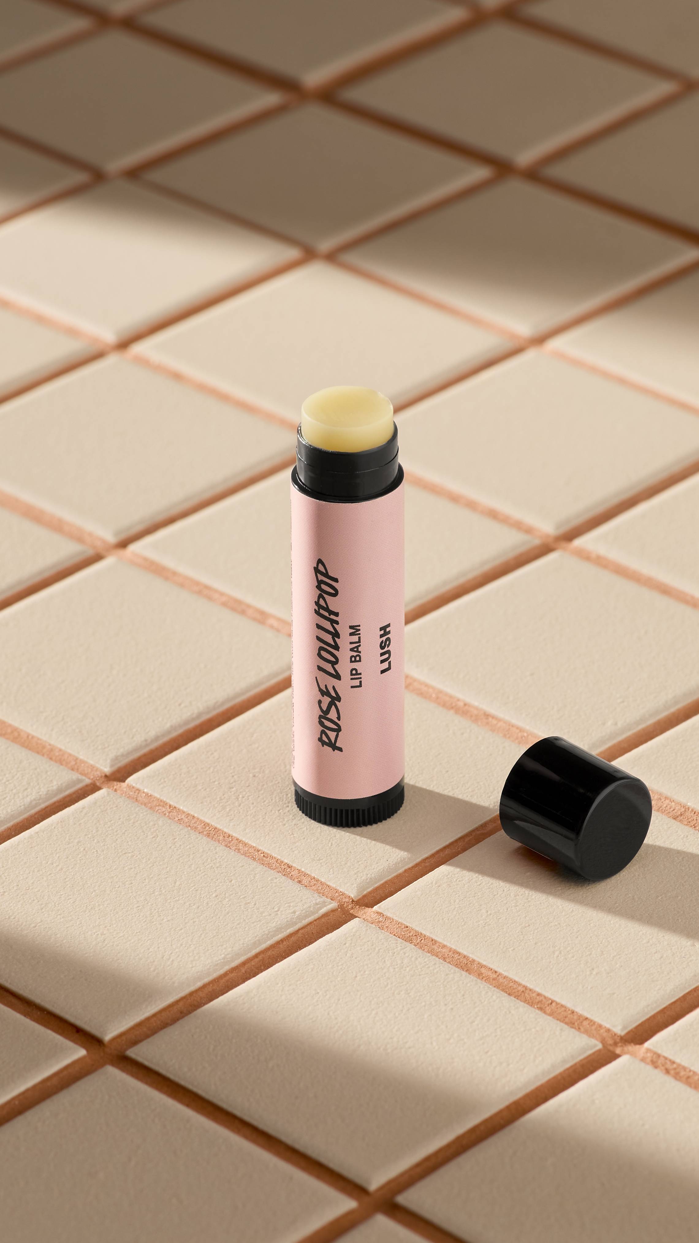 A light pink-labelled tube of Lush 'Rose Lollipop' lip balm stands upright on light-coloured square tiles. The black cap is off, lying next to the base, revealing the pale, creamy balm inside. Soft shadows fall across the tiled surface.