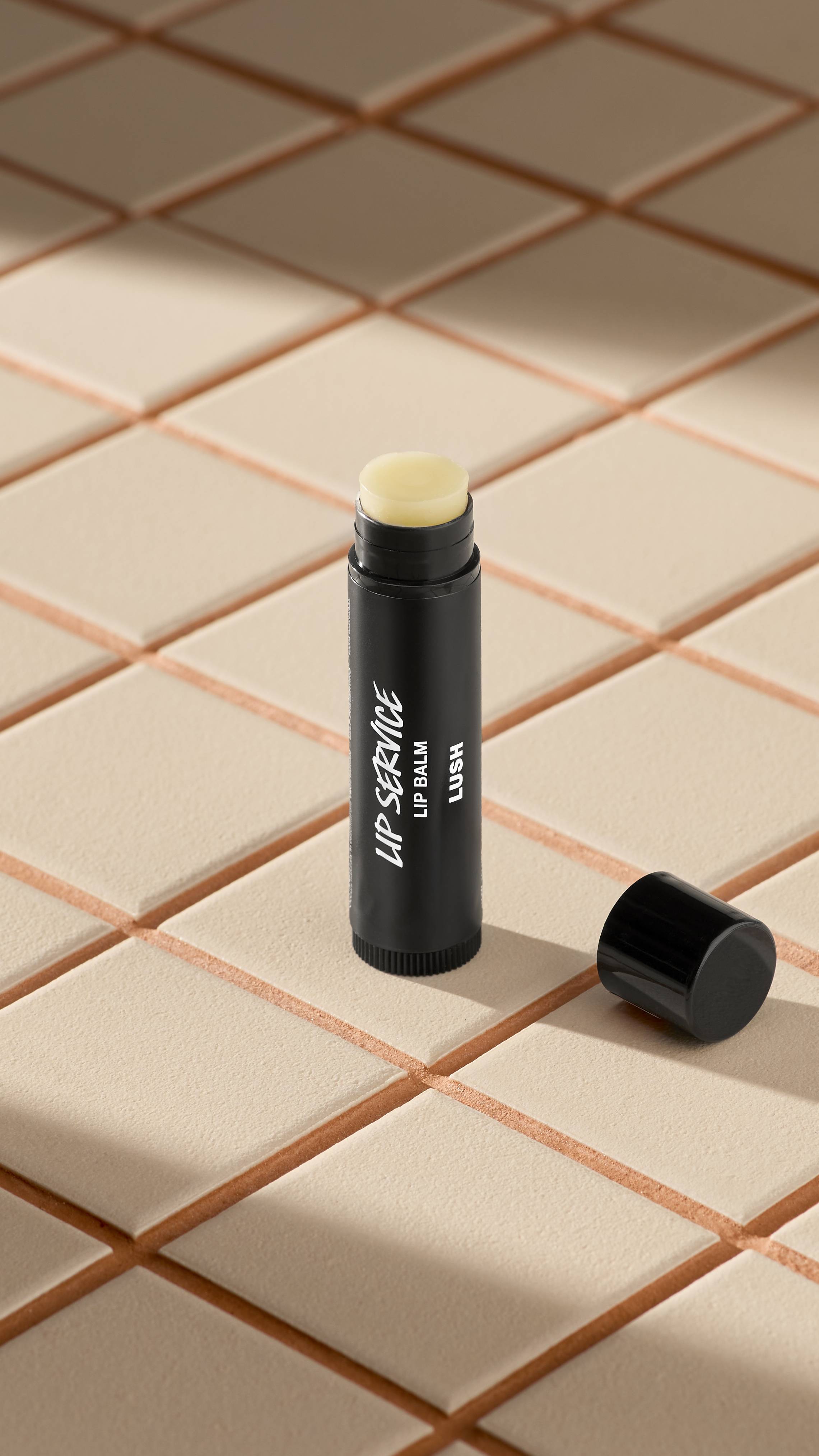 A black tube of Lush 'Lip Service' lip balm stands upright on light-coloured square tiles. The black cap is off, lying next to the base, revealing the pale, creamy balm inside. Soft shadows fall across the tiled surface.