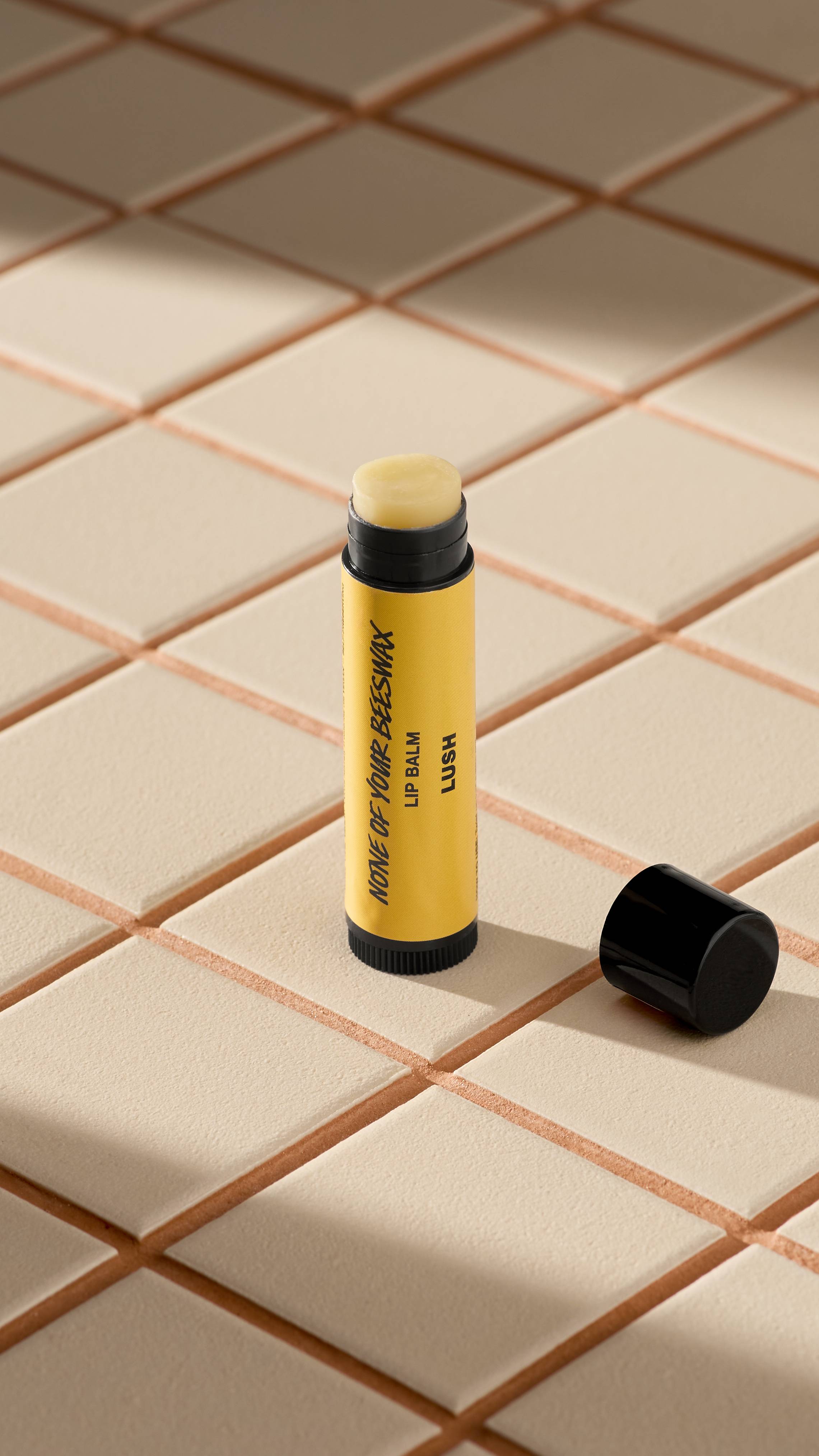 A yellow tube of Lush 'None of Your Beeswax' lip balm stands upright on light-coloured square tiles. The black cap is off, lying next to the base, revealing the pale, creamy balm inside. Soft shadows fall across the tiled surface.