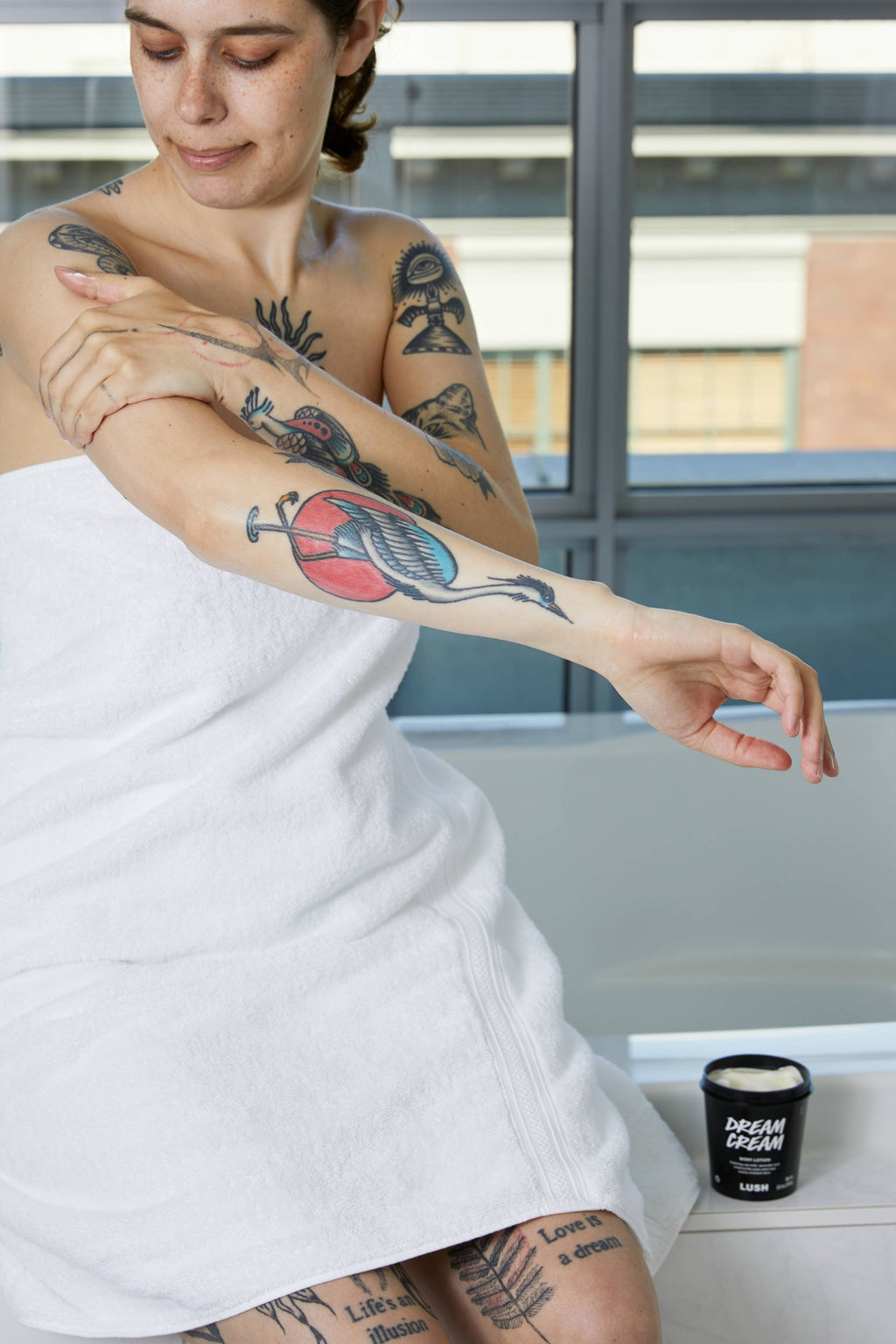 A tattooed person wrapped in a white towel sits on the edge of a bath, massaging cream into their arm. An open black tub of Lush 'Dream Cream' body lotion sits beside them on the tiled ledge.