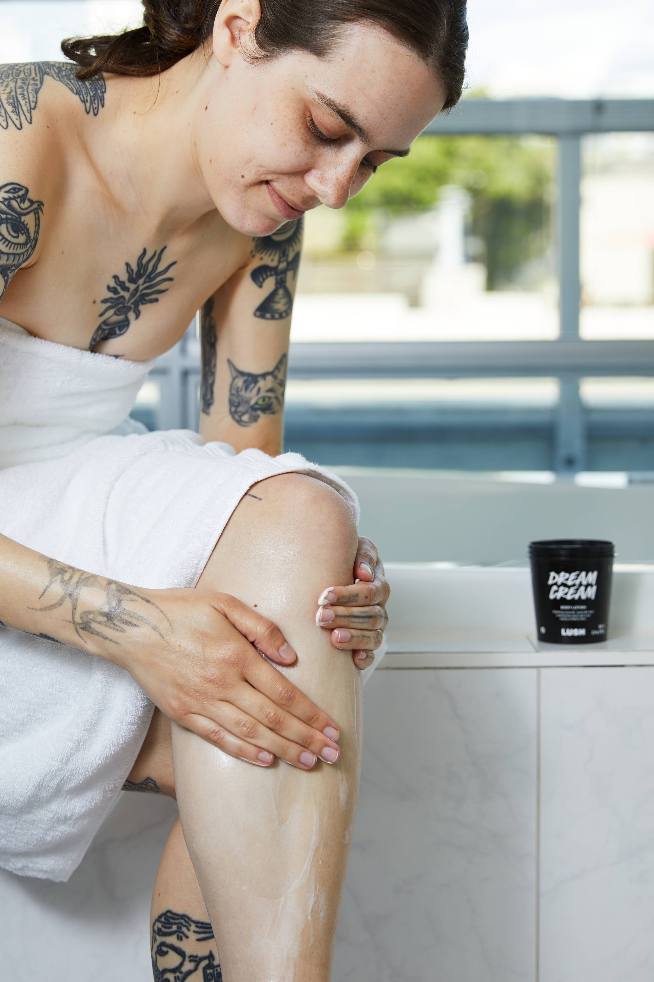 A tattooed person wrapped in a white towel sits on the edge of a bath, smoothing lotion onto their lower leg. In the background, a black tub of Lush 'Dream Cream' body lotion sits on the tiled ledge.