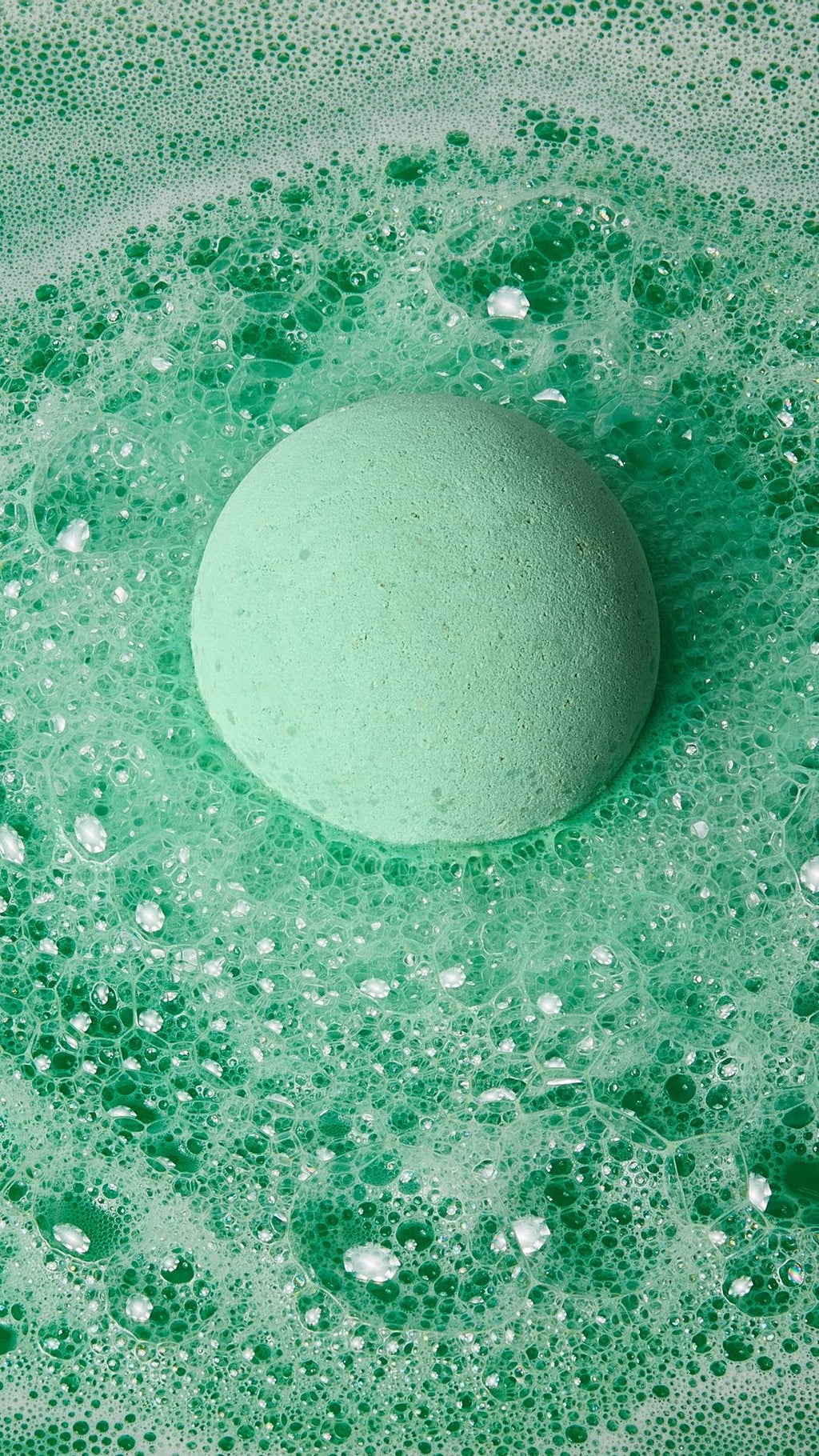 The image shows the pale green, round Avobath bath bomb floating on the surface of vibrant green bath water. Thick, foamy bubbles surround the bomb, creating a swirling texture across the water.