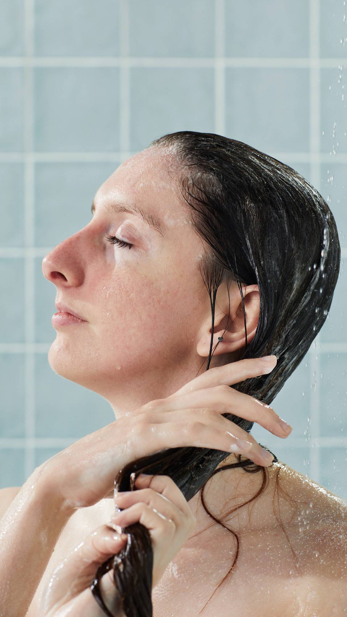 A side-profile view of a model with eyes closed, running the white Lush "American Cream" conditioner through their long, dark hair in the shower. Their hands are coated in product against a backdrop of glossy, light blue square tiles.