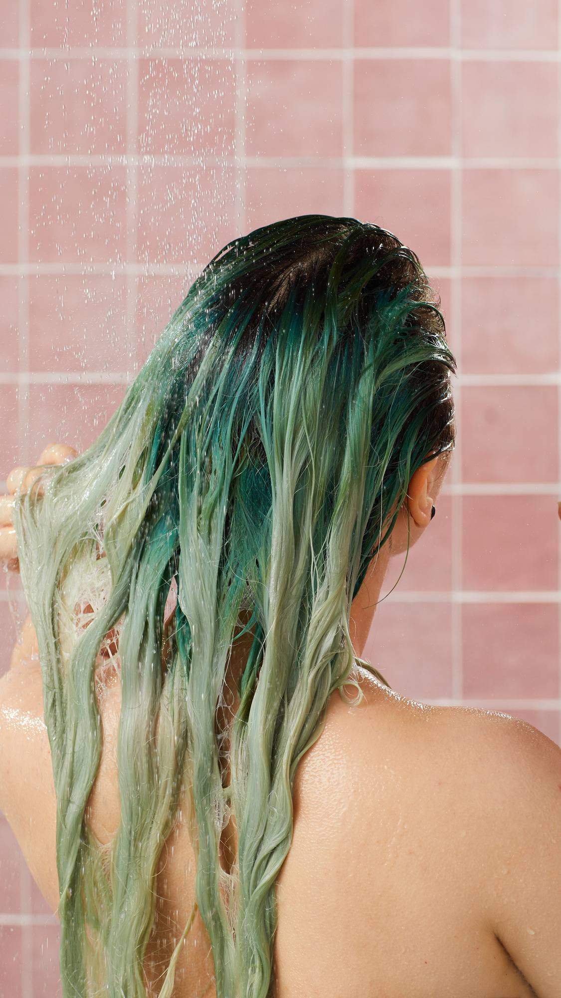 A view from behind of a person with long, wet hair dyed in shades of green and teal as they wash it under a running shower, after using Lush "Retread" conditioner. Water cascades over their hair and shoulders against a backdrop of pink square tiles.