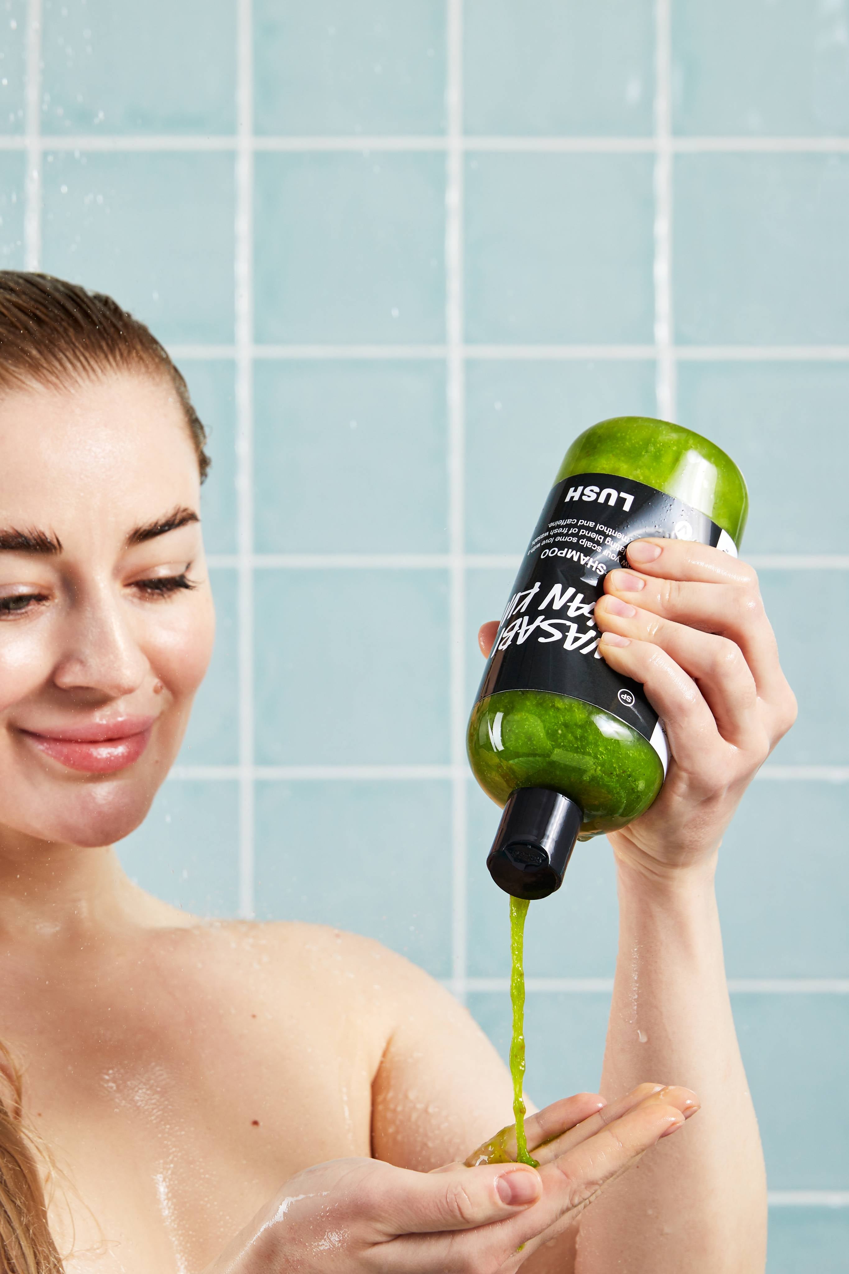 A smiling person in the shower squeezes an upside-down bottle of green Lush Wasabi Shan Kui shampoo, pouring a stream of textured green liquid into their hand. Set against a background of pale blue square wall tiles.