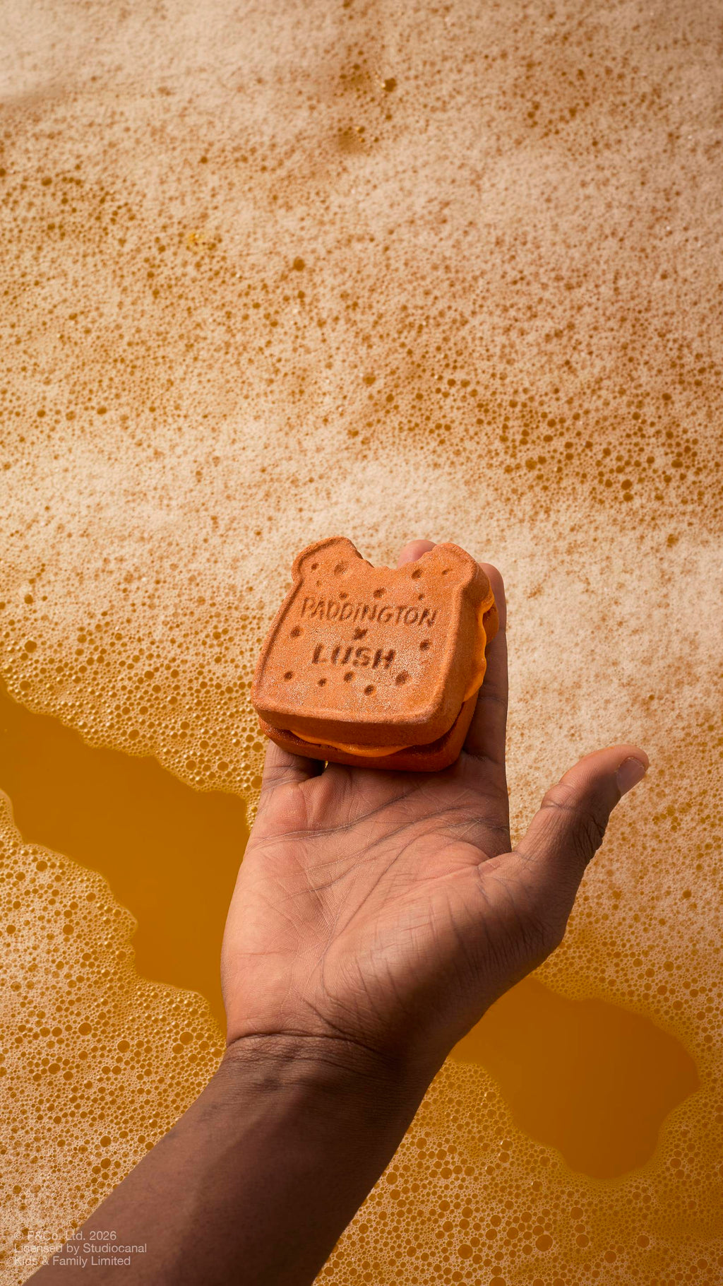 A model's hand holds a "Marmalade Sandwich" bubbleroon bubble bar over golden, foamy bathwater. The brown, textured bubble bar features the embossed text 'PADDINGTON x LUSH' and has a small 'bite' taken out of the top.