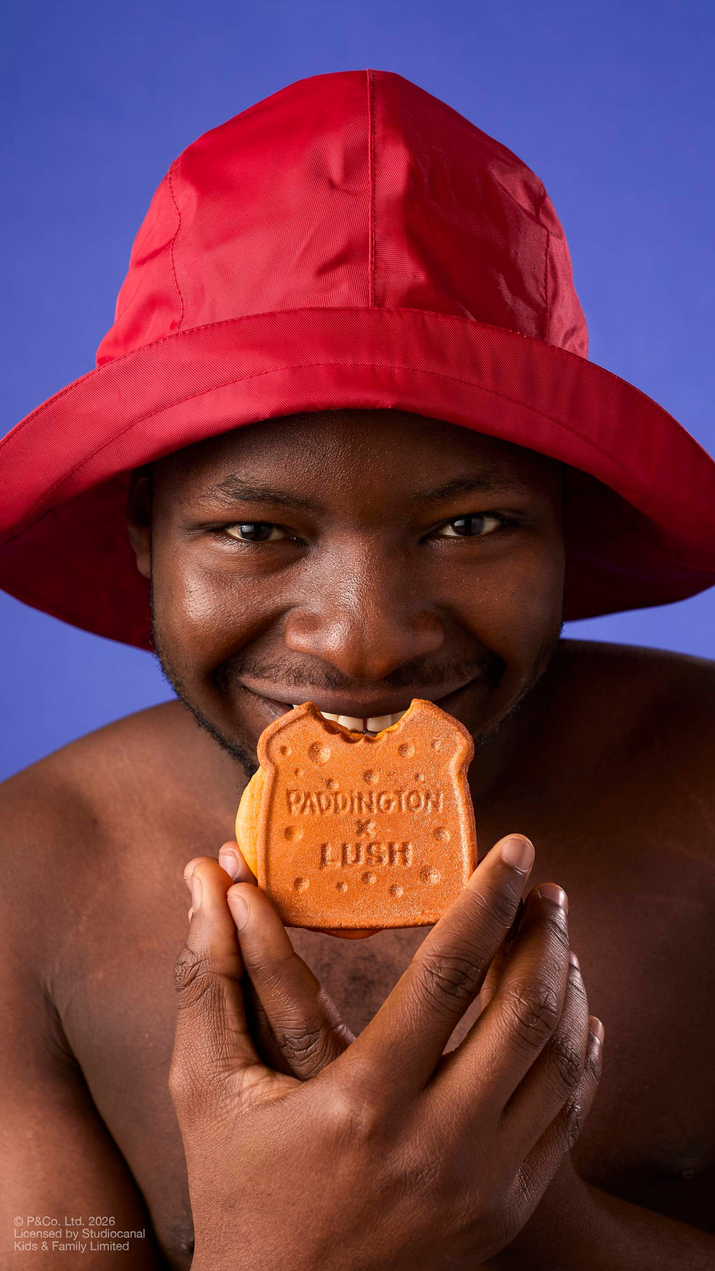 A model in a red rain hat smiles directly at the camera while holding a "Marmalade Sandwich" bubbleroon bubble bar to their mouth, as if about to take a bite. The bubble bar features the text 'PADDINGTON x LUSH' against a solid purple background.
