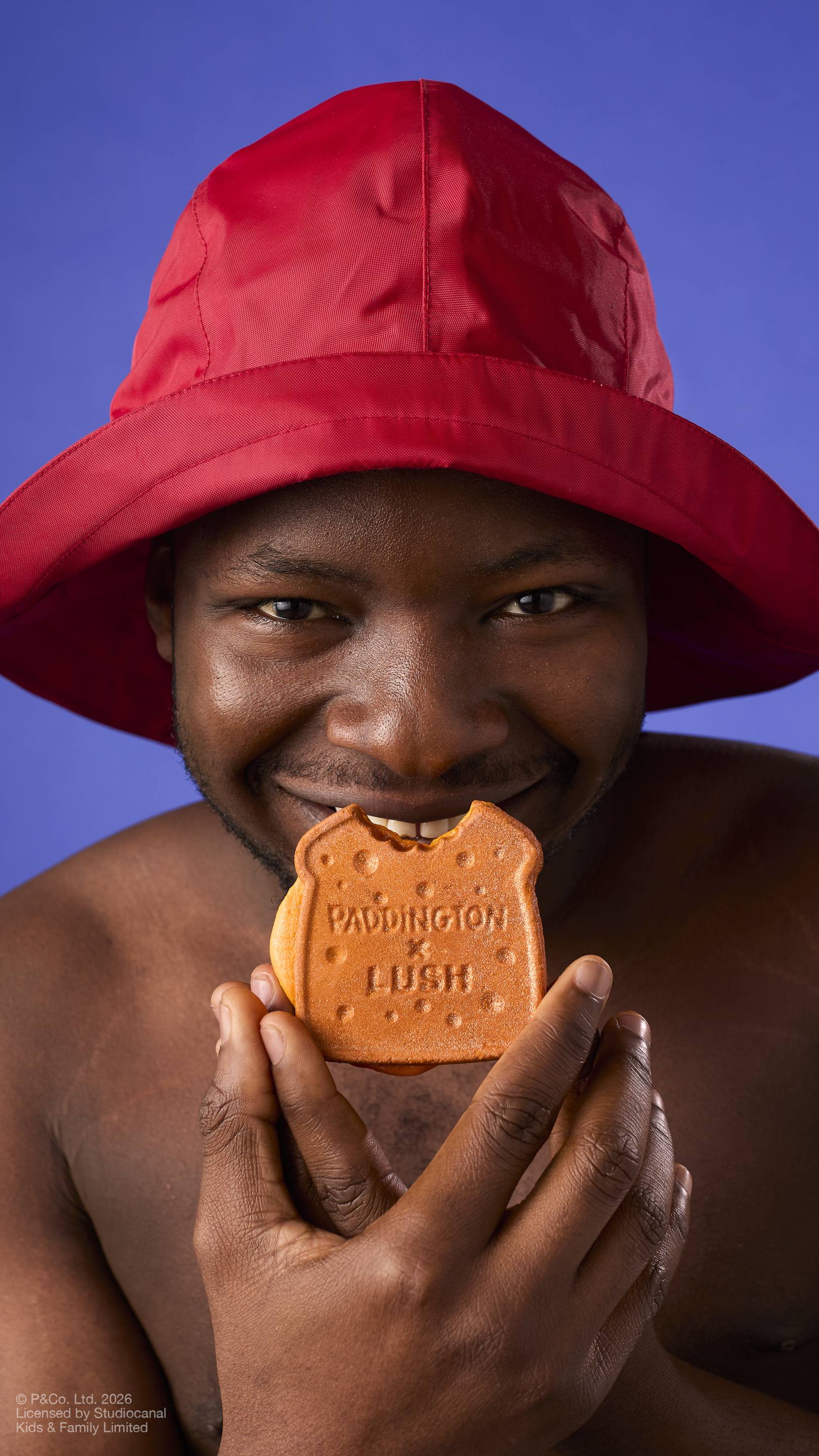 A model in a red rain hat smiles directly at the camera while holding a "Marmalade Sandwich" bubbleroon bubble bar to their mouth, as if about to take a bite. The bubble bar features the text 'PADDINGTON x LUSH' against a solid purple background.