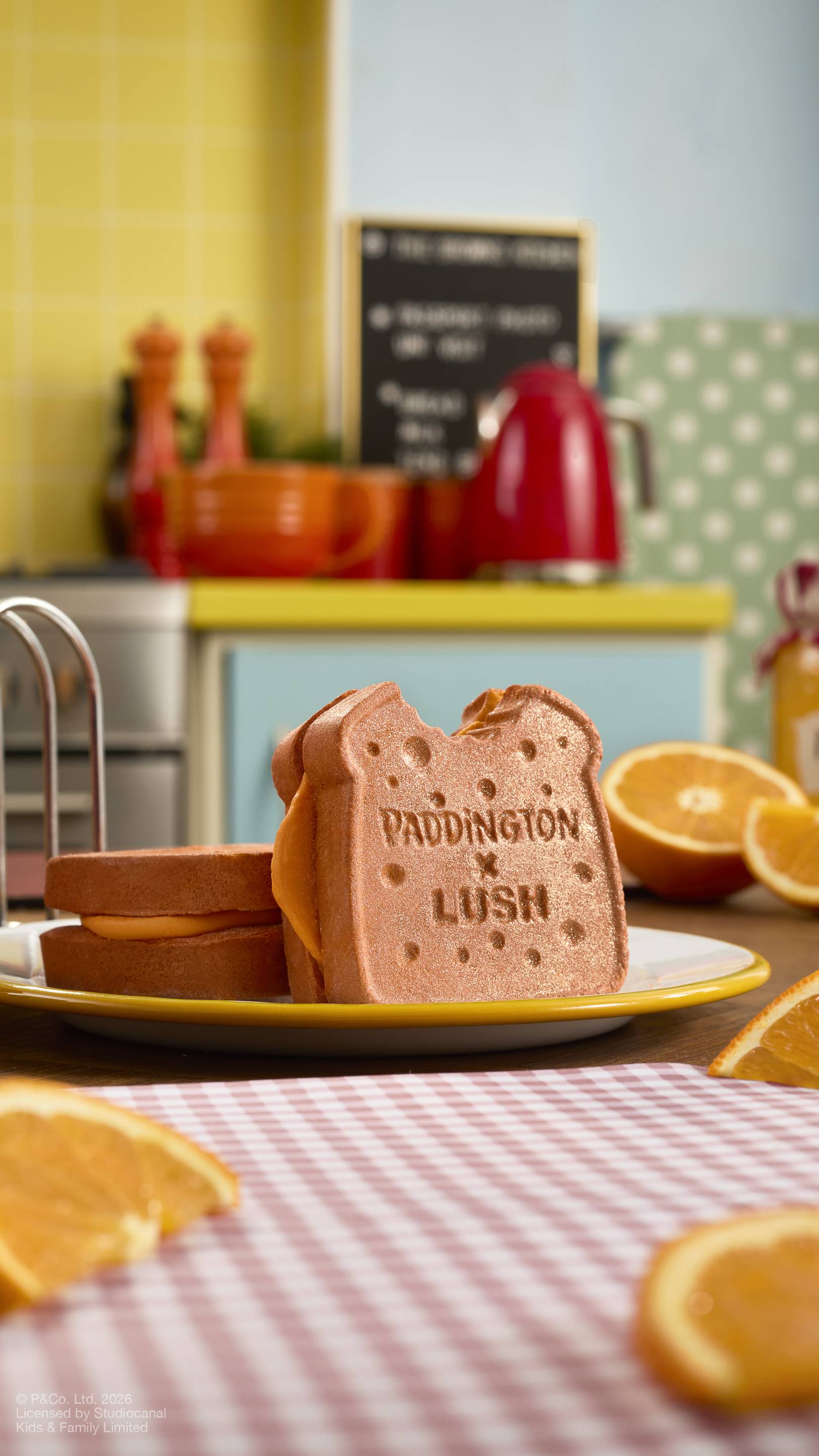 The image shows two brown, "Marmalade Sandwich" bubbleroon bubble bars sitting on a plate on a kitchen counter. One stands upright, showing a 'bite' taken out of the corner and 'PADDINGTON x LUSH' text. Surrounded by orange slices.