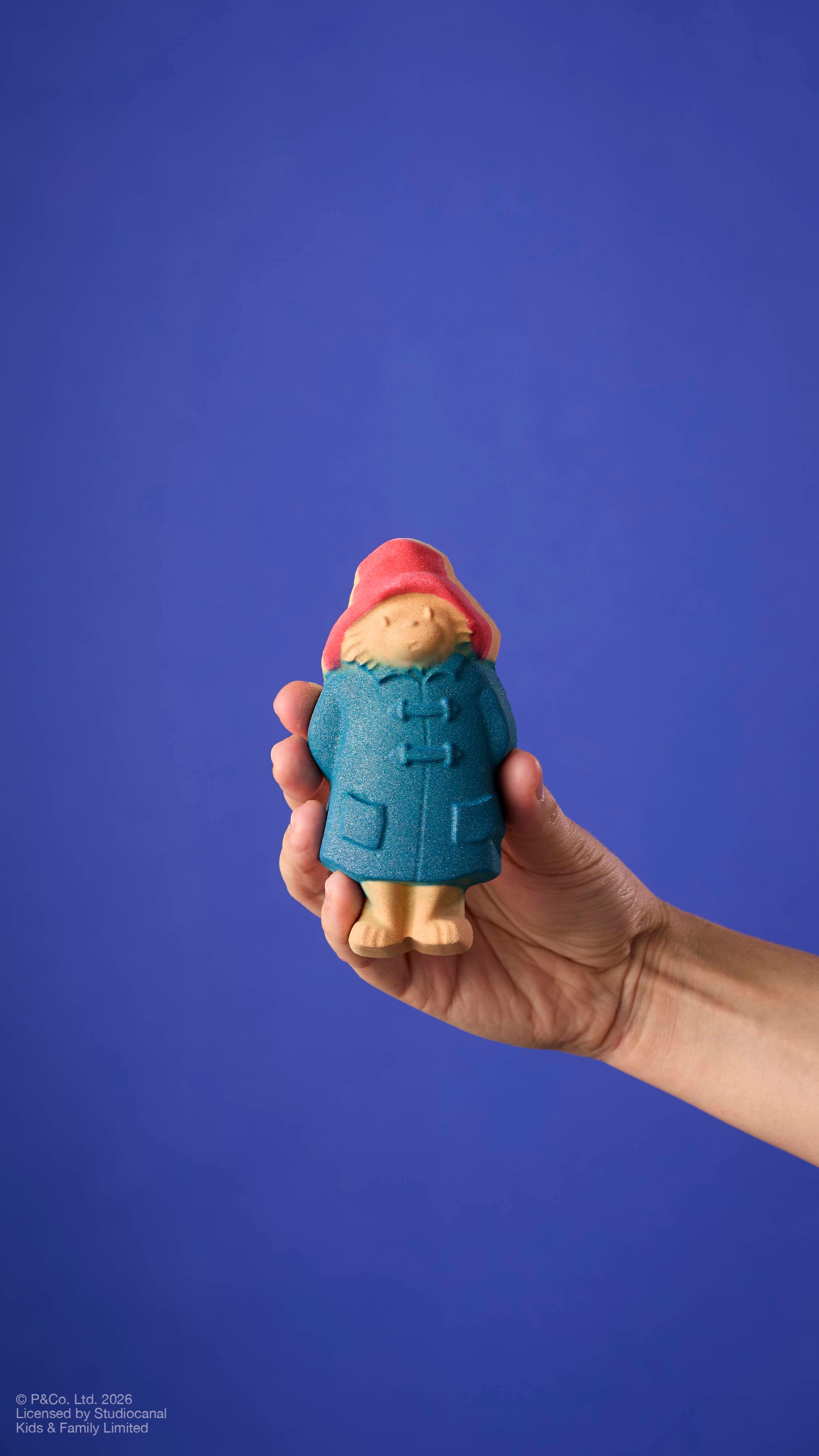 A model's arm holds a "Paddington Bear" bath bomb against a solid blue background. The bear wears his signature red hat and blue duffle coat with textured button details.
