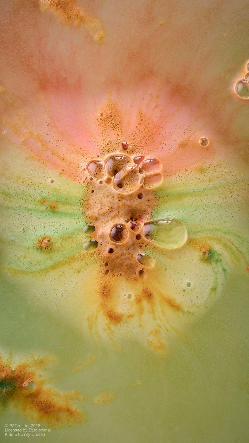 An aerial shot of bath water as the 'Paddington Bear" bath bomb dissolves, creating a marbled effect. Thick, frothy bubbles in shades of amber, yellow and green congregate in the centre, surrounded by swirling patterns.
