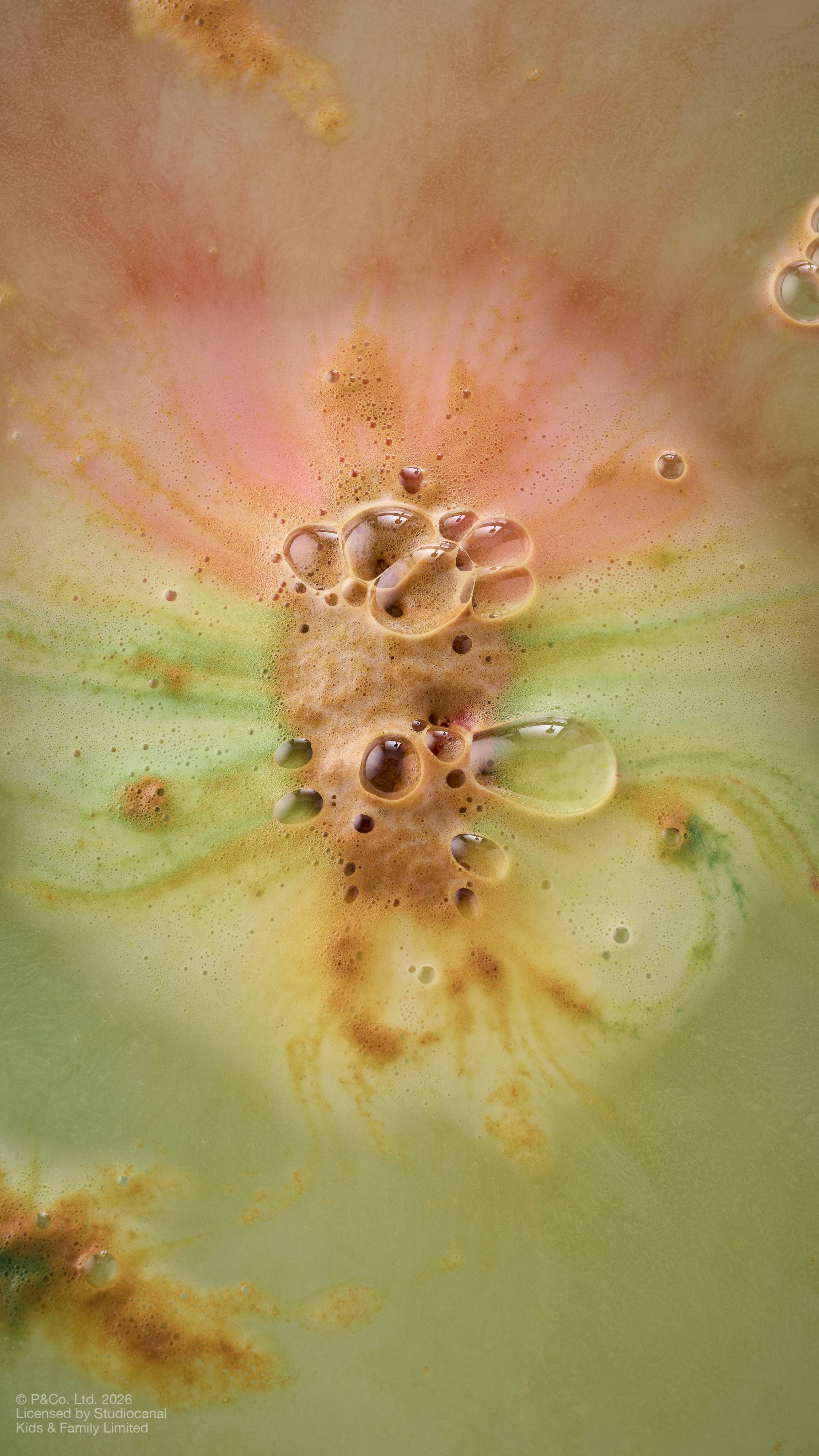 An aerial shot of bath water as the 'Paddington Bear" bath bomb dissolves, creating a marbled effect. Thick, frothy bubbles in shades of amber, yellow and green congregate in the centre, surrounded by swirling patterns.